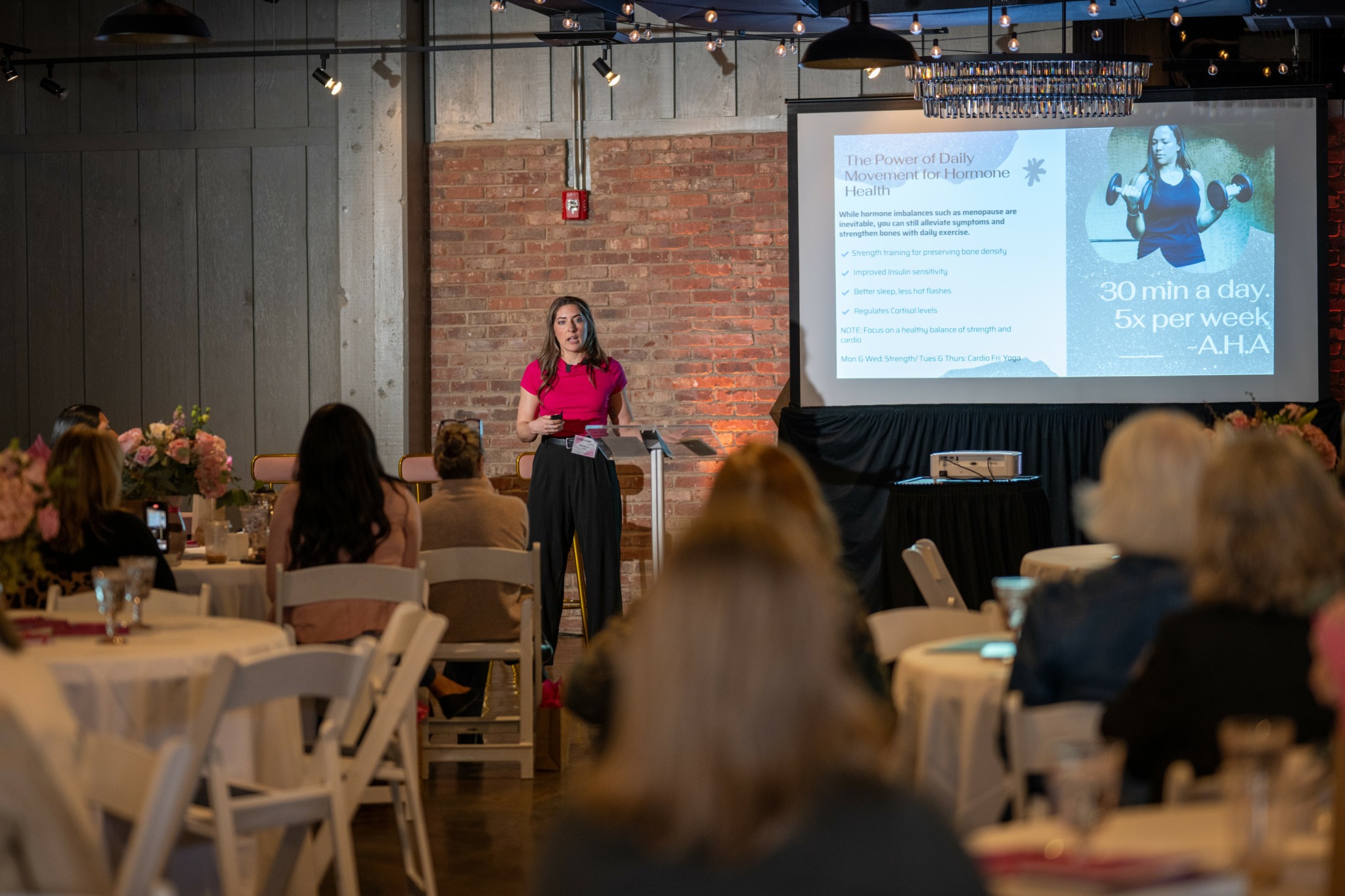 Woman presenting in front of audience with a slide projected on screen, brick wall background, indoor event setting.
