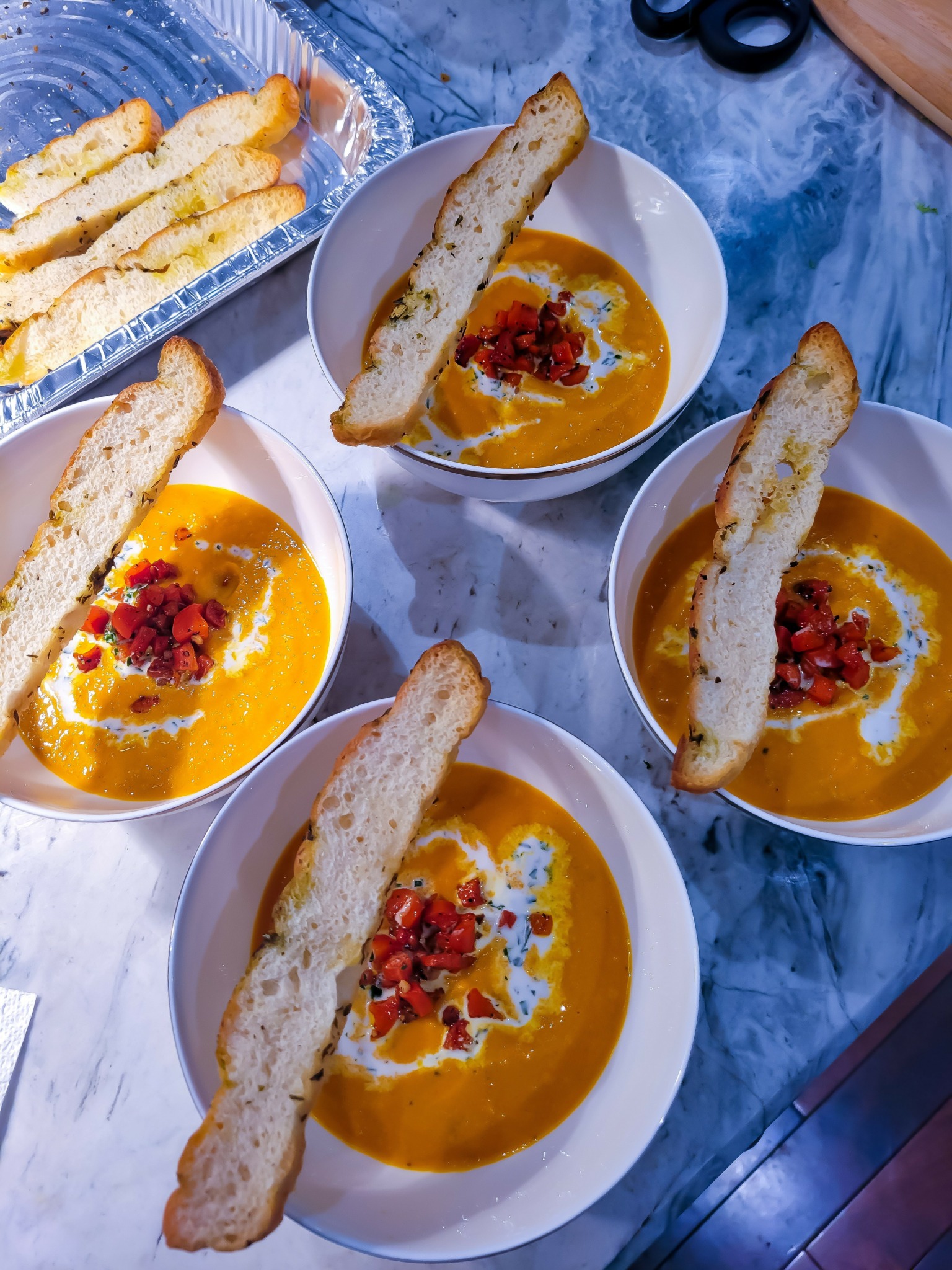 Four bowls of creamy orange soup topped with red garnish and bread slices on a marble surface.
