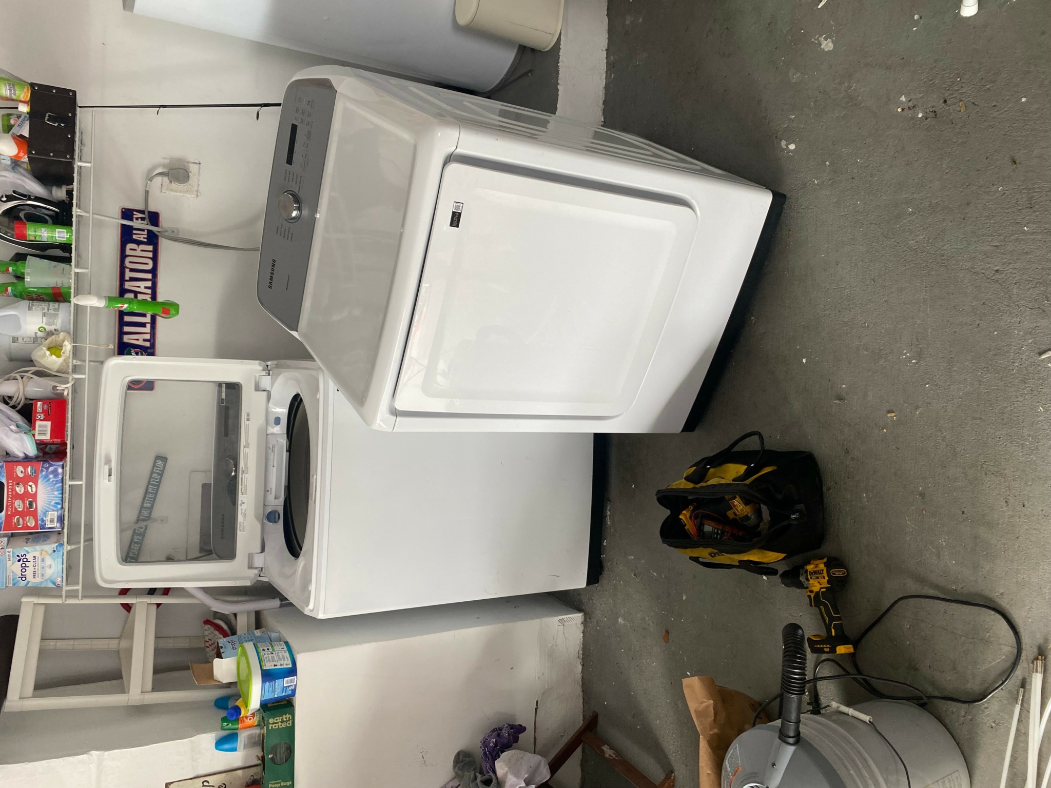 Front view of a washing machine and dryer in a laundry room with cleaning supplies and tools nearby.