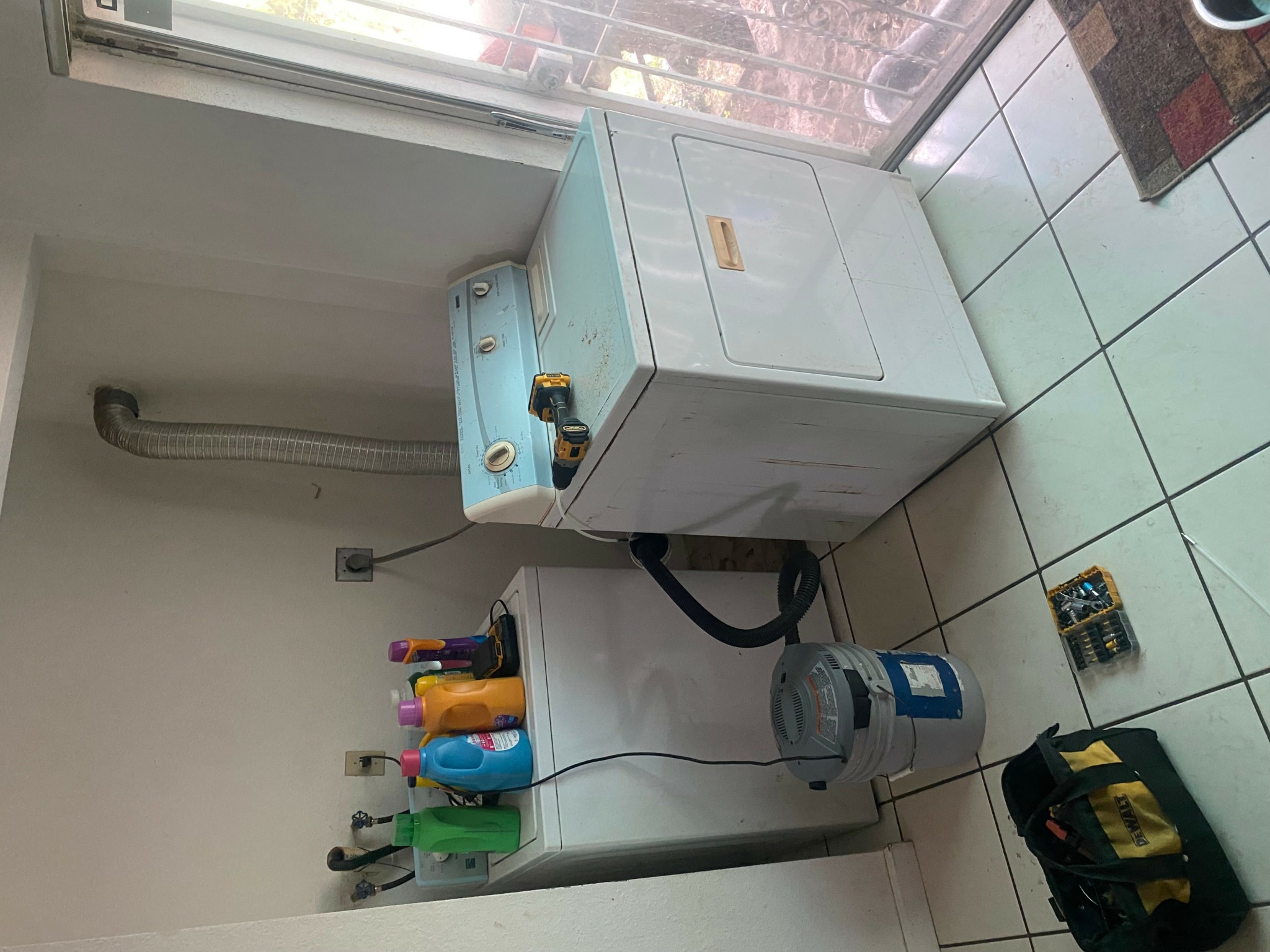 Washer with laundry supplies on top, hoses connected, and a water filter on tiled floor, wall, and window in background.