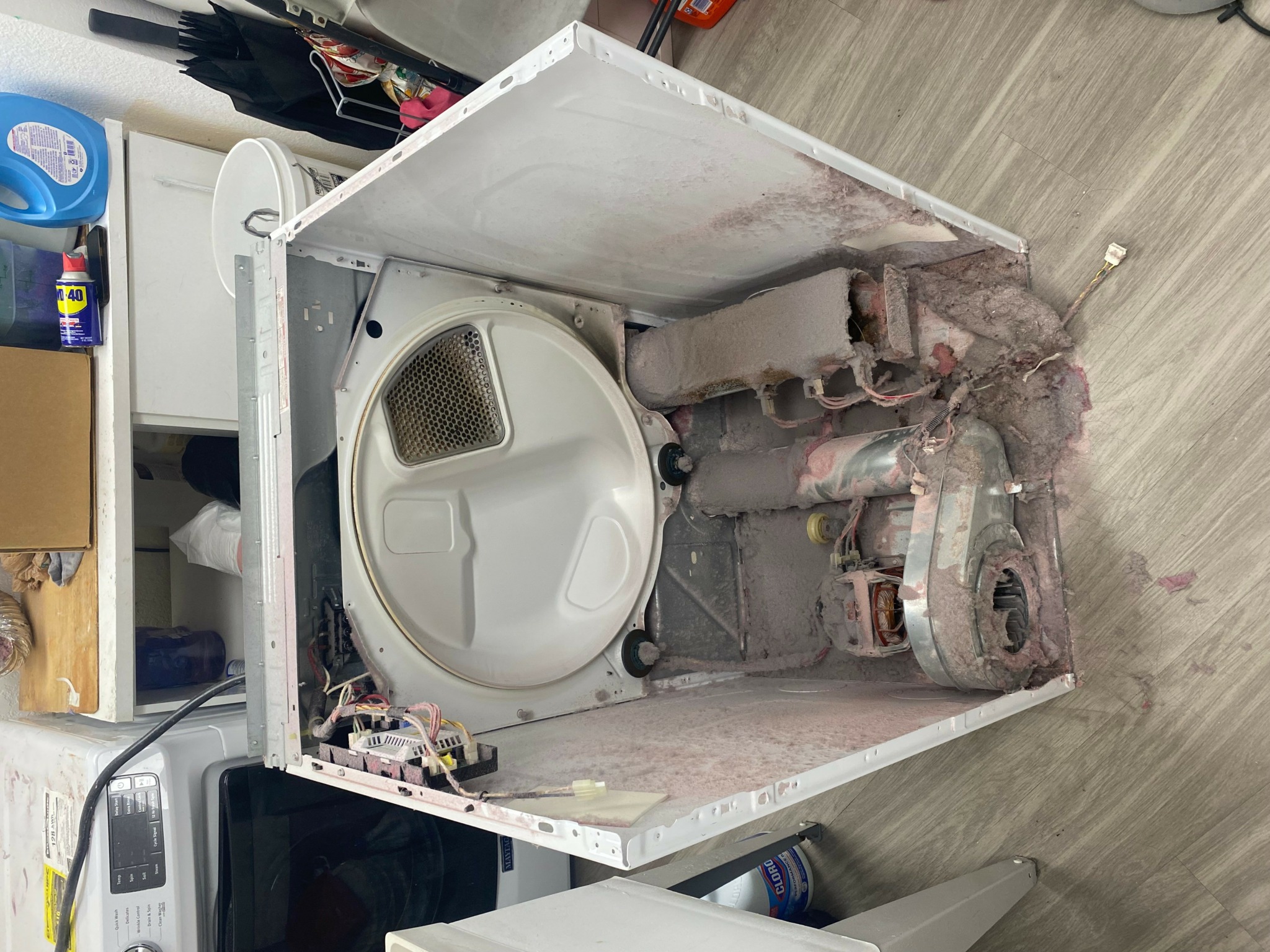 Open washing machine with drum visible inside, placed inside a small, white, partially enclosed space, with a wooden floor nearby.