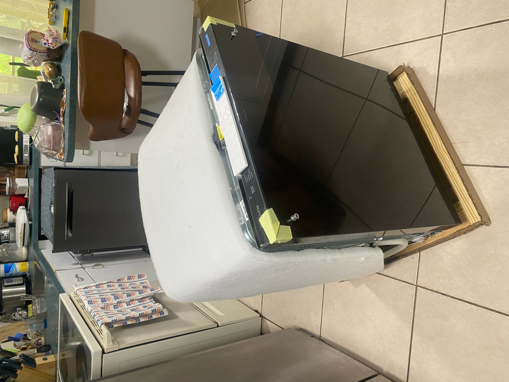 Flat-screen TV on a wooden stand, wrapped in protective foam, in a room with tiled floor and various objects in background.