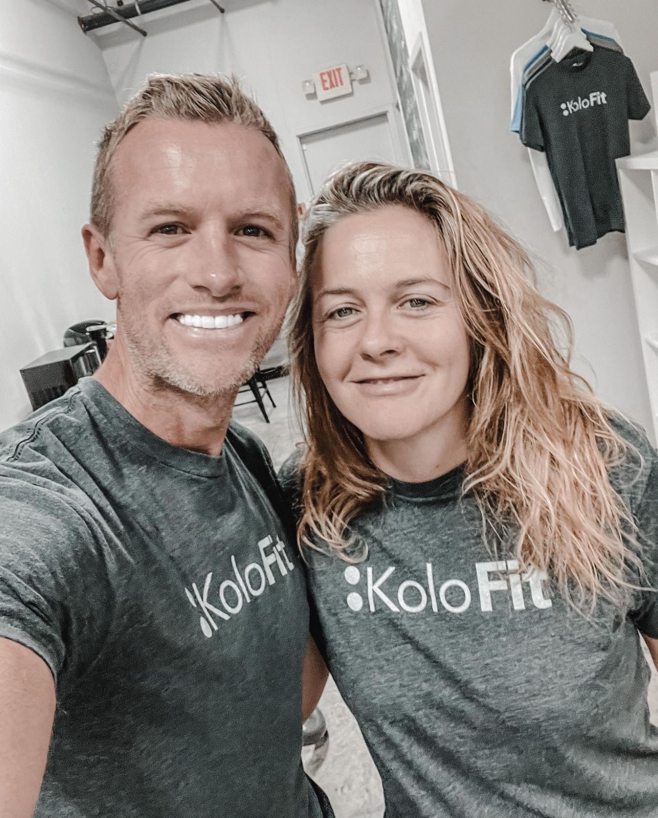 Two people smiling, wearing KoloFit shirts, standing indoors with gym equipment and clothing in the background.