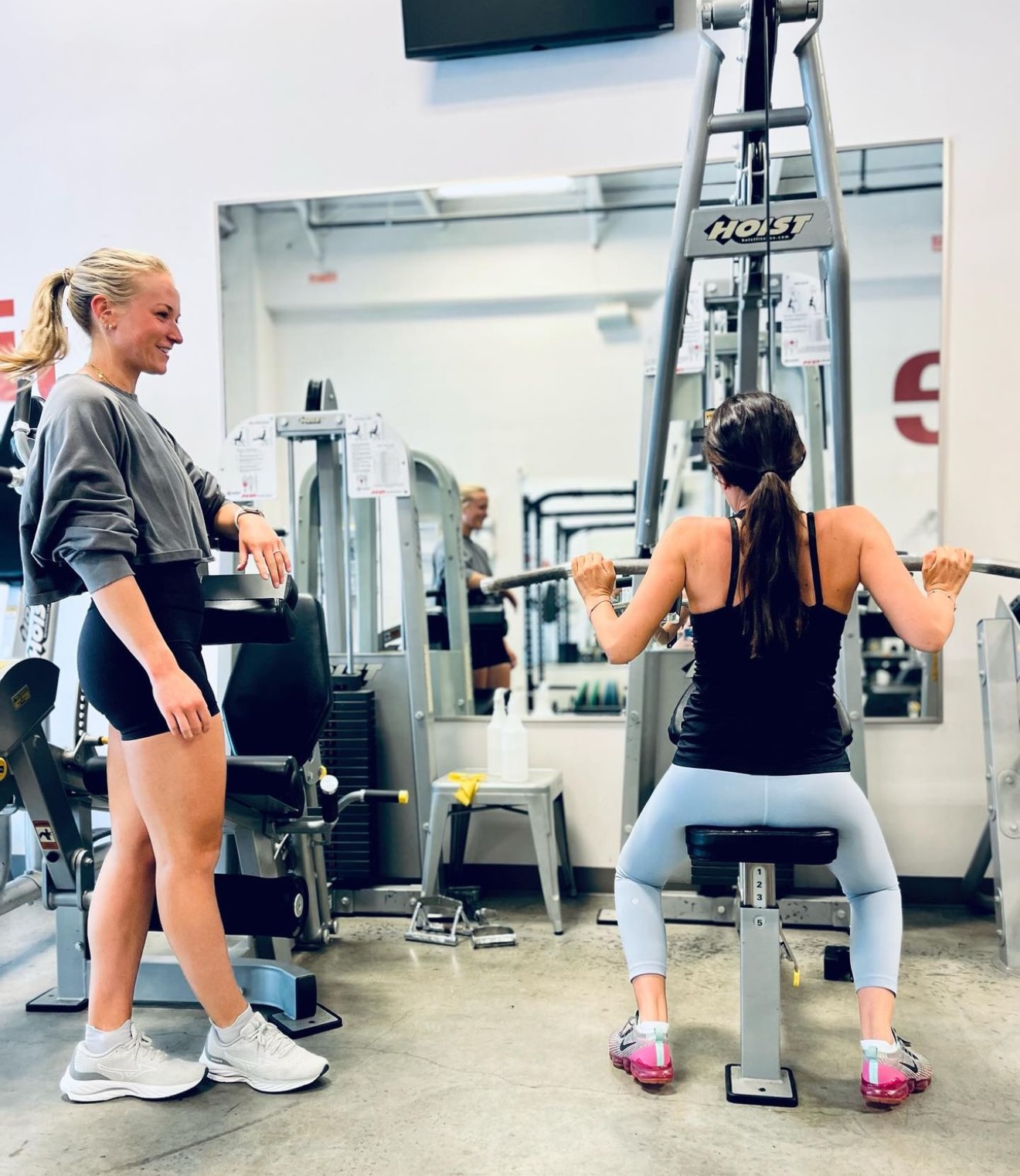 Two women exercising in a gym, one sitting on a machine and the other standing nearby, with gym equipment in the background.