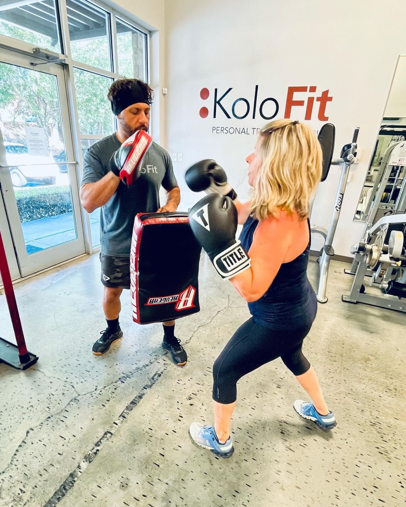 Woman in boxing gloves practicing punches with trainer in gym, large window and fitness equipment in background.
