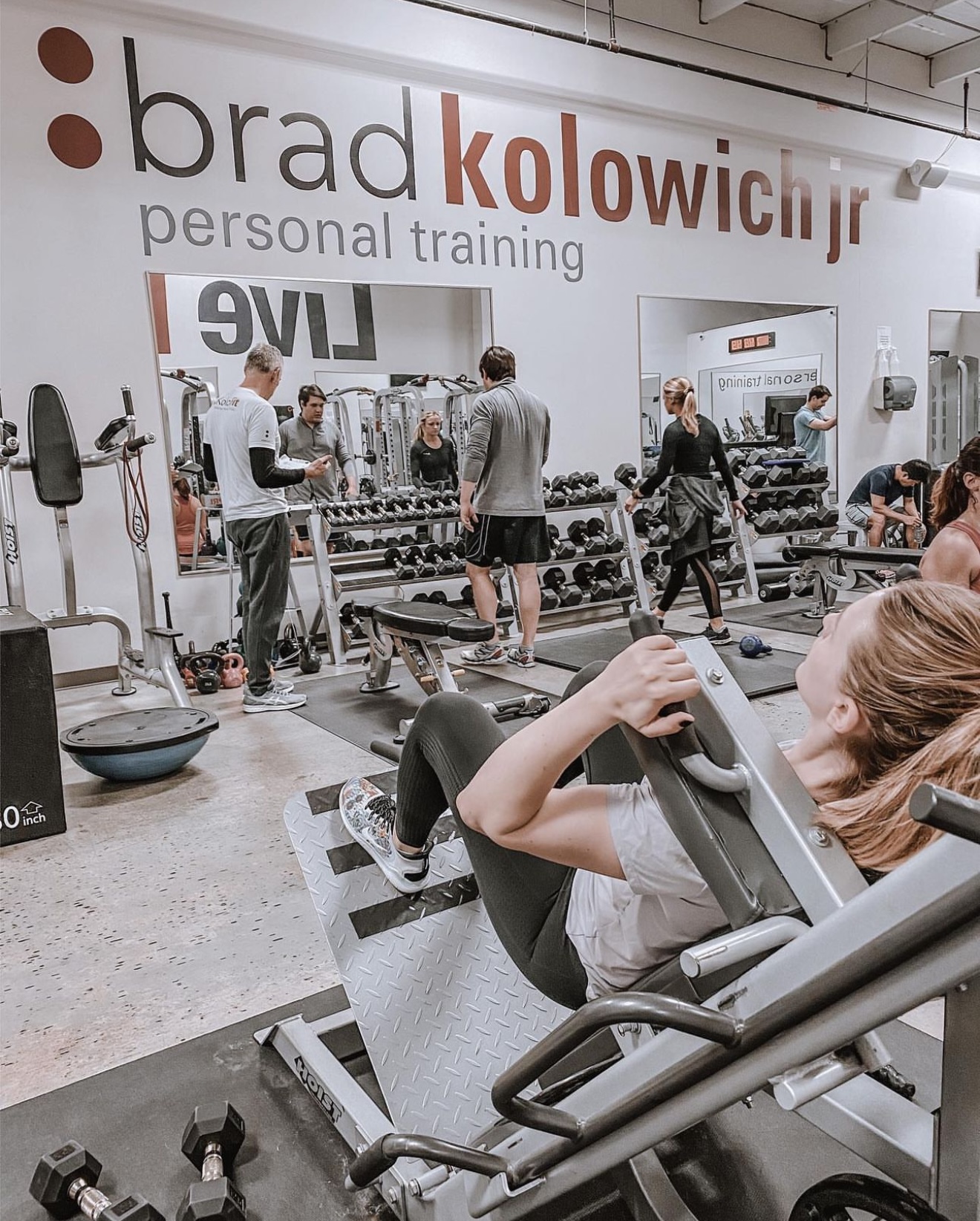 People exercising with weights and machines in a gym, with a large wall sign reading 'brad kolowich jr personal training'.