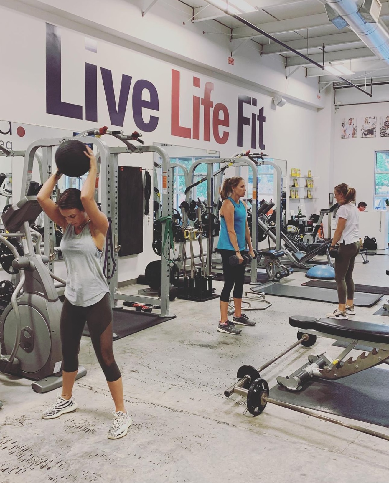 People exercising with weights and equipment in a gym with a large 'Live Life Fit' sign on the wall.