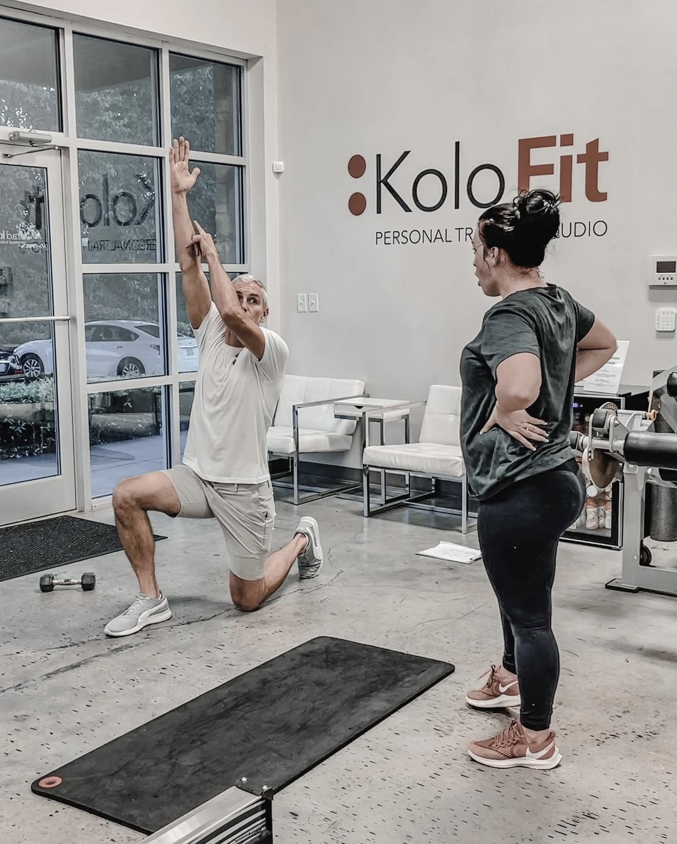 Person performing a knee exercise with arms raised, trainer observing in a gym with fitness equipment and a logo on the wall.