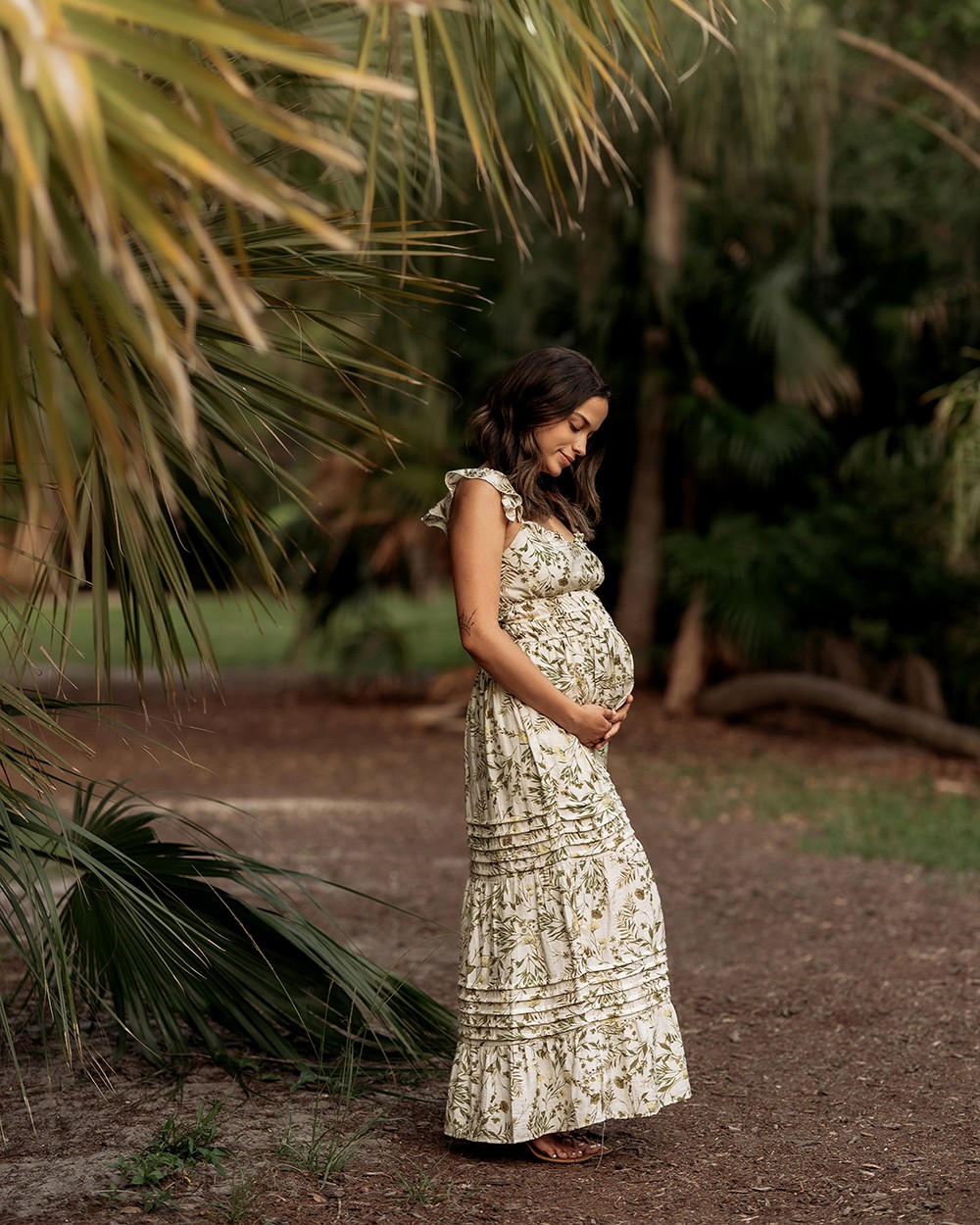 Pregnant woman standing outdoors, holding her belly, surrounded by greenery and trees, smiling gently, wearing a long patterned dress.