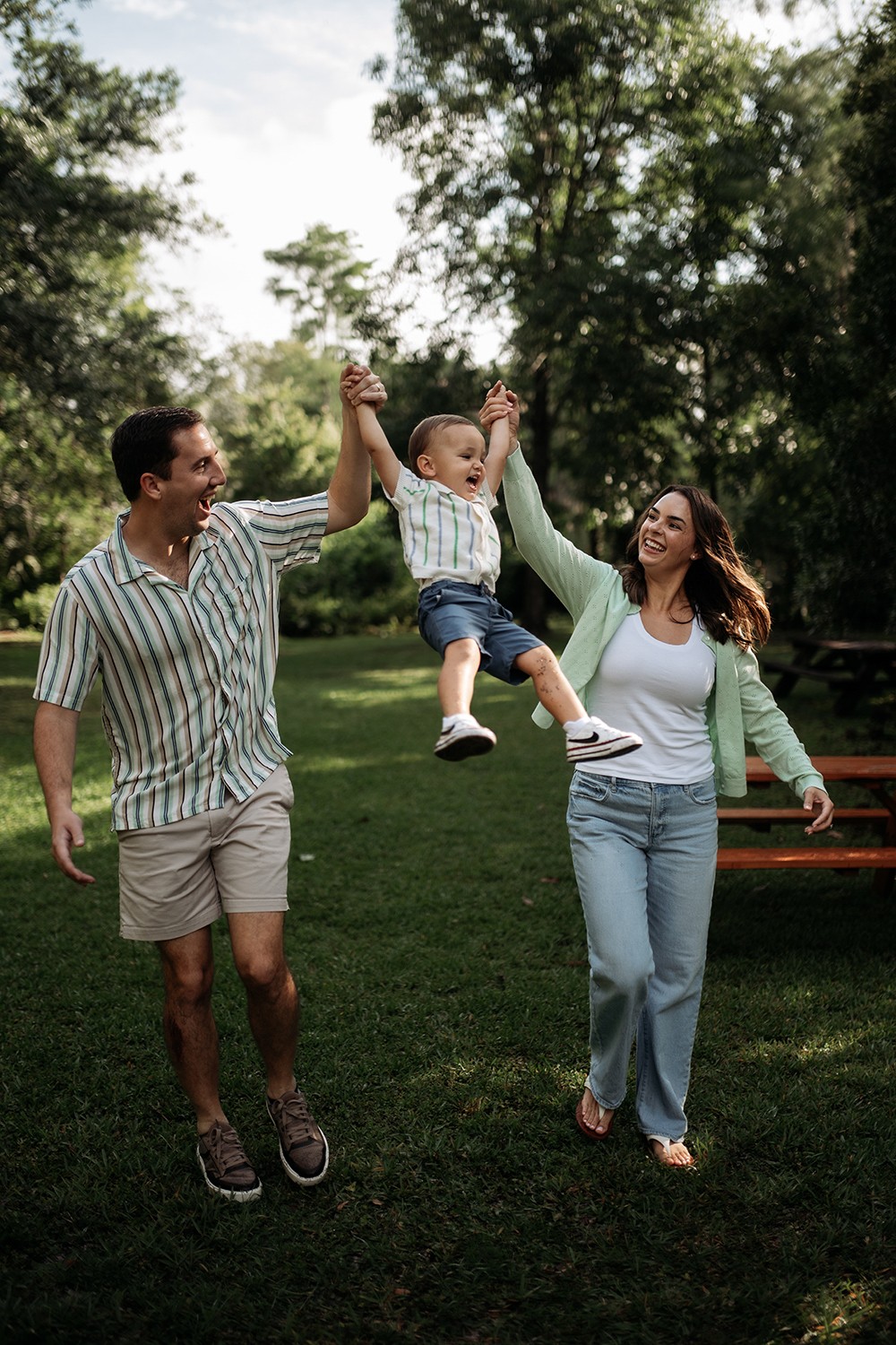 Family of three outdoors, father and mother holding child's hands, child swinging in the air, trees in background