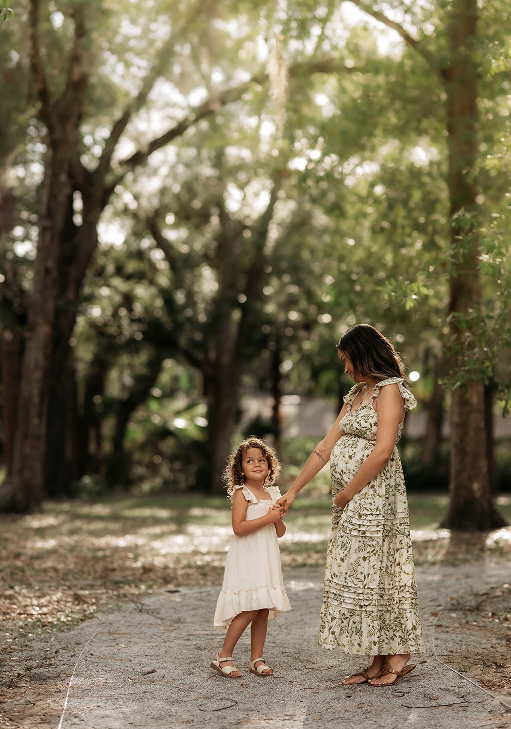 Woman and young girl holding hands in a wooded park, sunlight filtering through trees, girl in white dress, woman in patterned dress