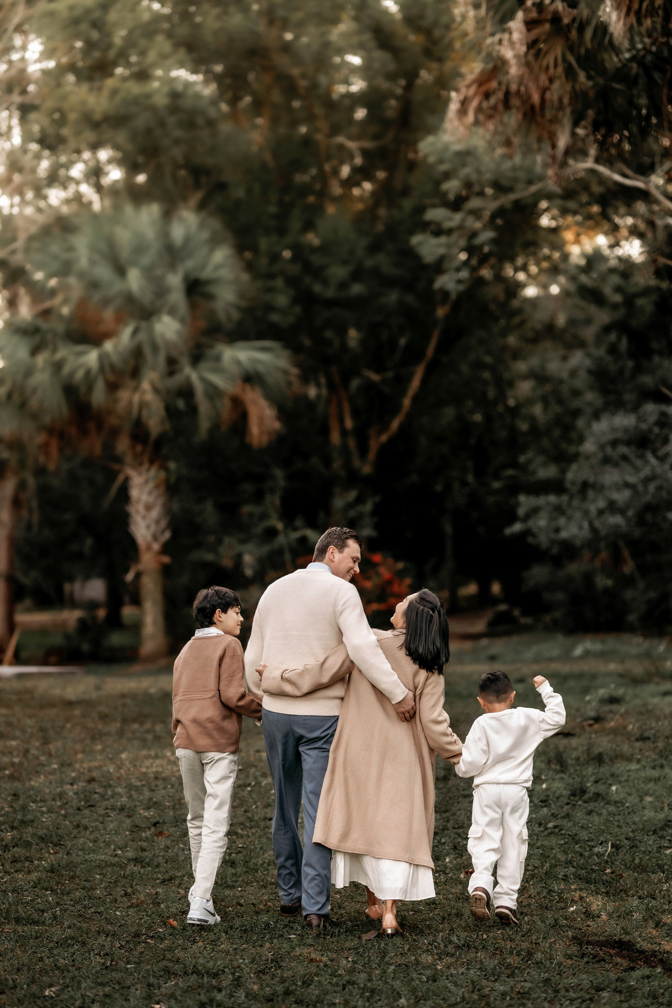 A family of four walking in a park with trees and greenery, holding hands and enjoying the outdoors.