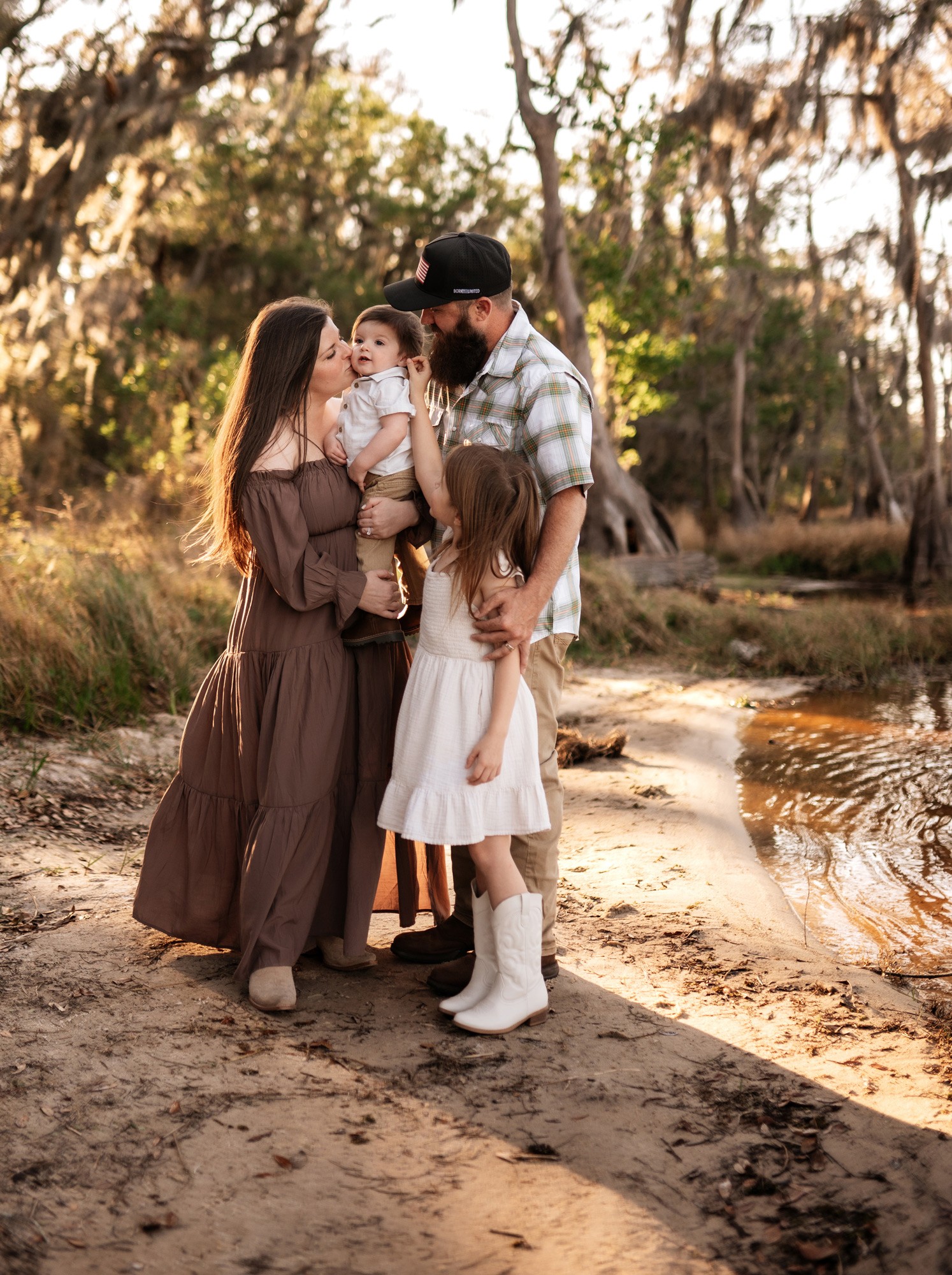 Family of four standing outdoors near water, with trees and sunlight in background.