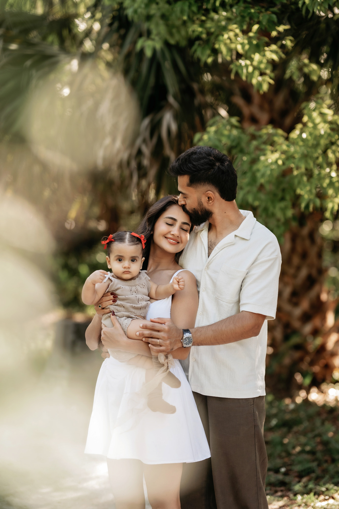 Family of three outdoors, woman holding a child, man leaning close, surrounded by greenery.