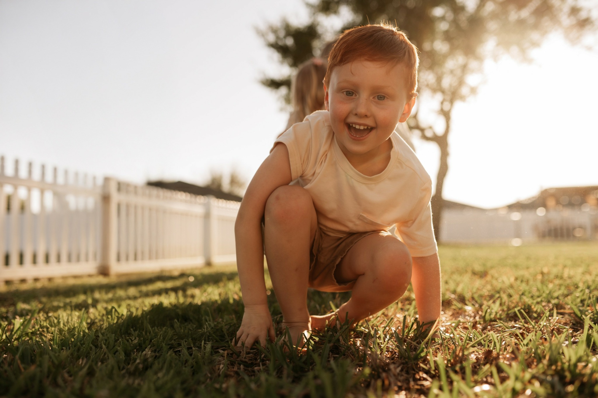 Child crouching on grass outdoors, smiling, with a white fence and trees in the background, sunlight shining through.