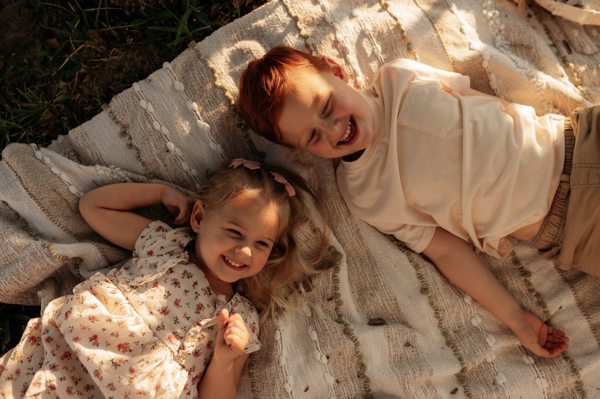 Two children lying on a blanket outdoors, smiling and laughing, with sunlight shining on them.