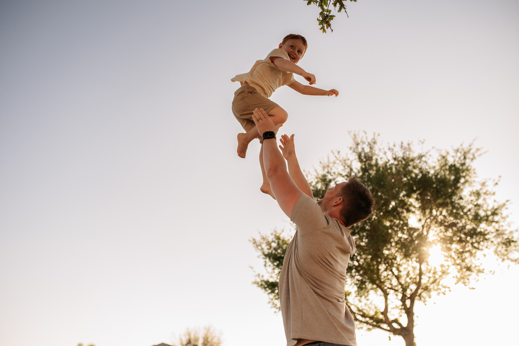 A man throws a child into the air outdoors with trees and sky in the background.