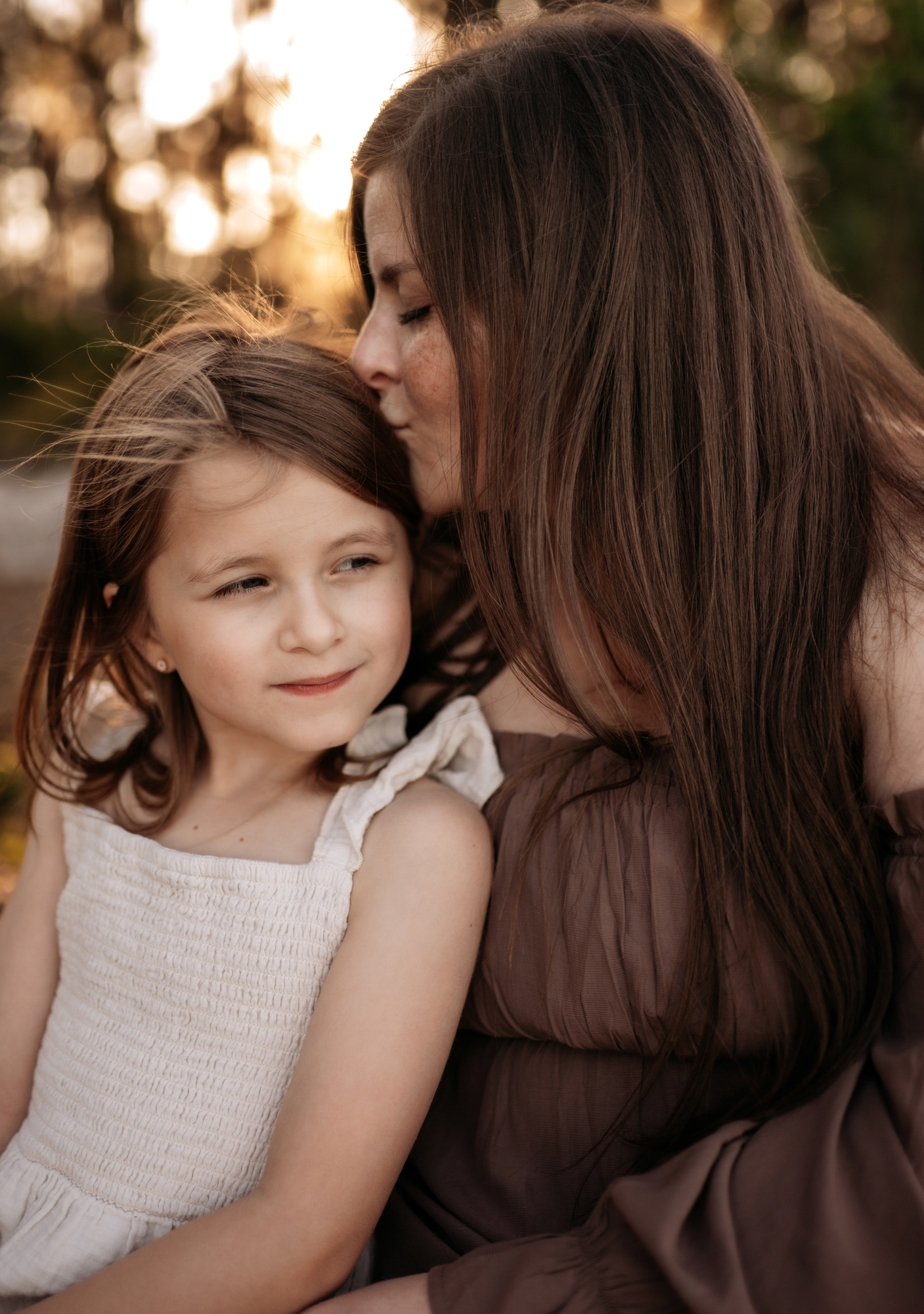 Woman and girl close together outdoors, woman gently touching girl's head, warm sunlight background, natural setting