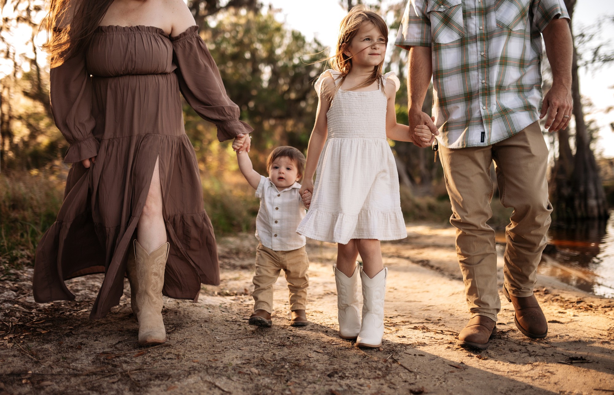 Family walking outdoors on a dirt path, holding hands, with trees in the background, during sunset.