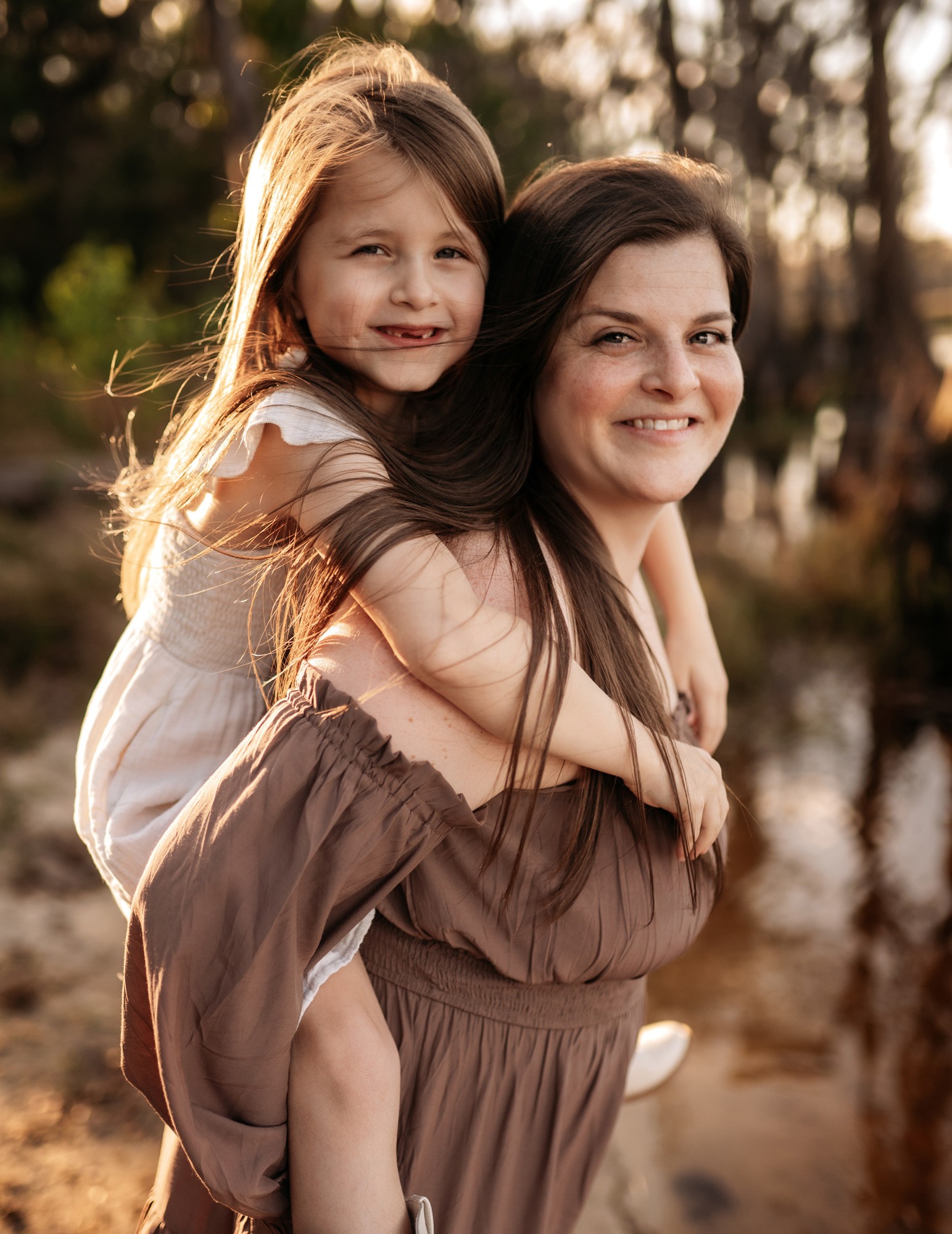 A woman carries a young girl on her back outdoors, both smiling, with trees and water in the background.