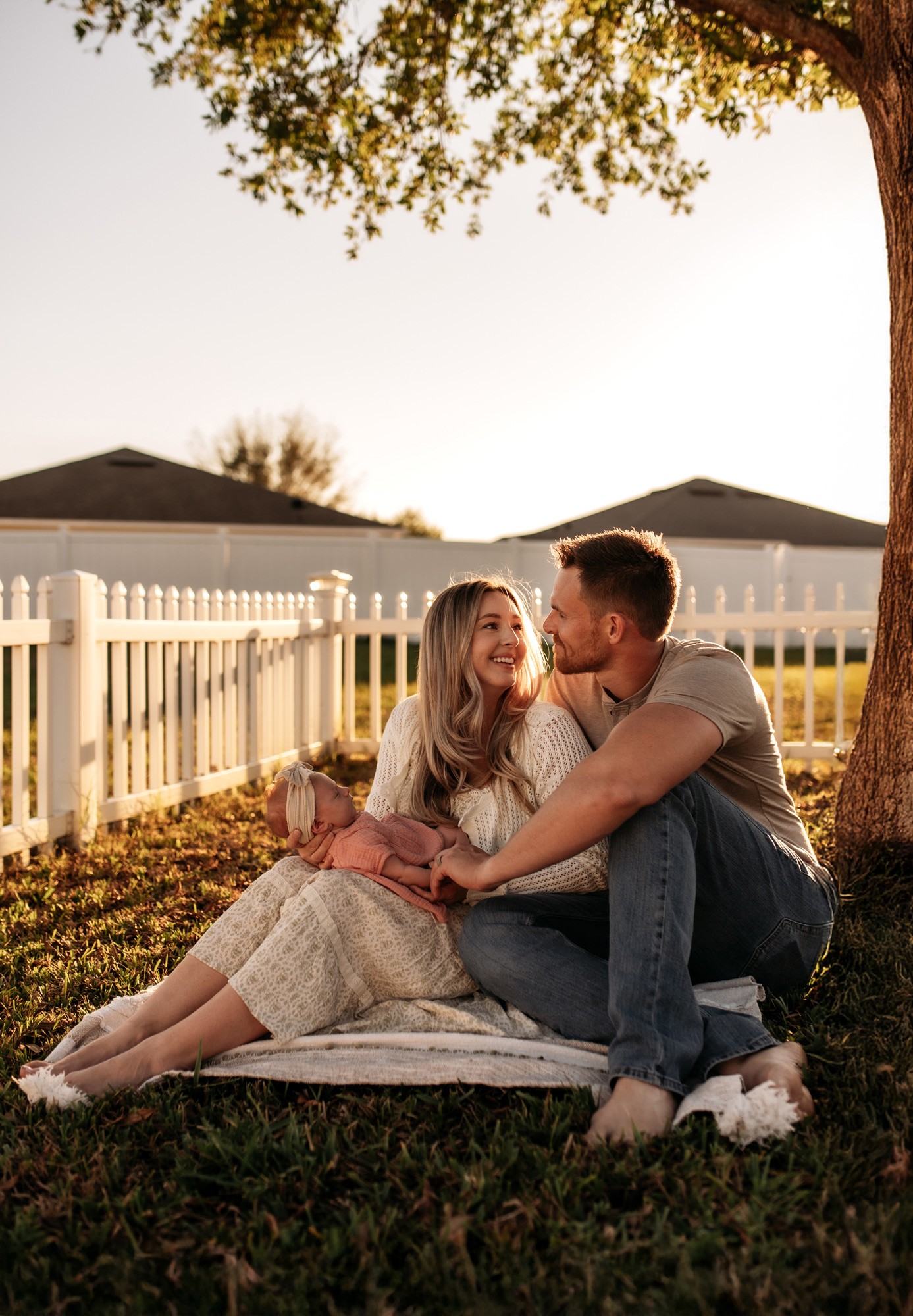 A couple sitting on a blanket under a tree in a backyard, smiling at each other, with a white fence and houses in background.