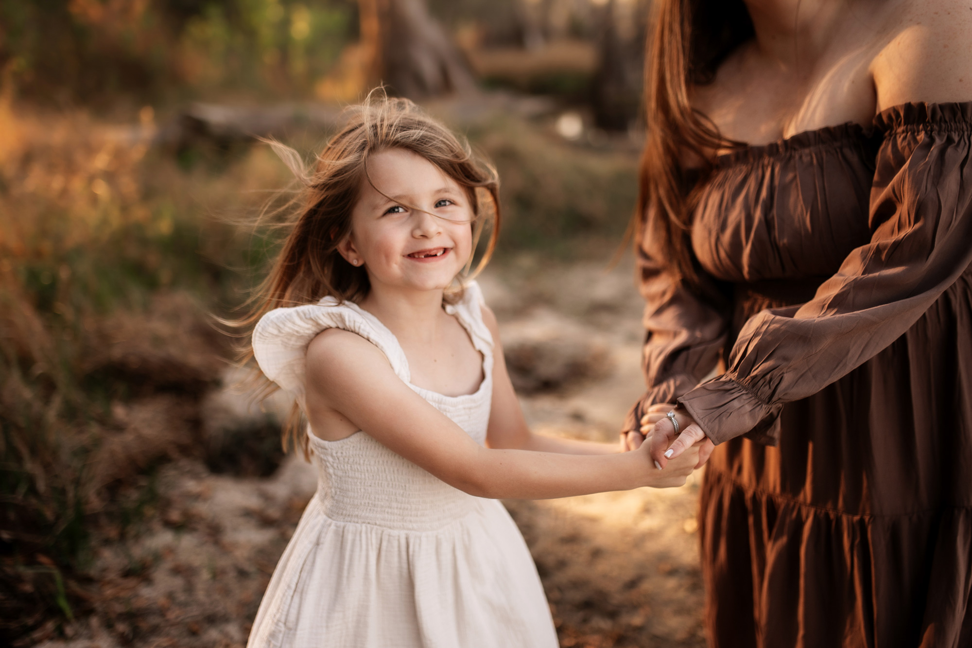 Young girl with long hair smiling and holding hands with an adult, outdoors in a natural setting.