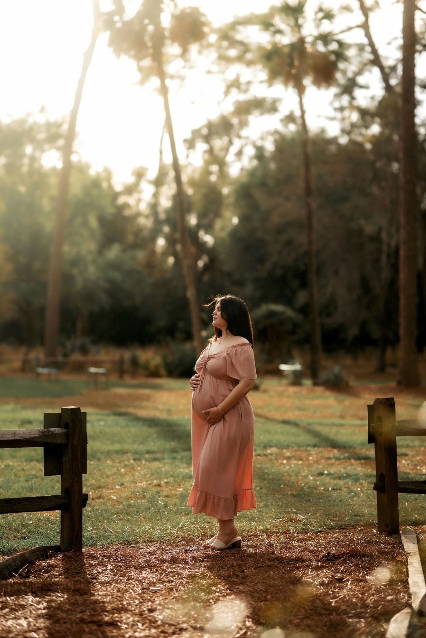 Pregnant woman in pink dress standing outdoors near a wooden fence, trees, and sunlight filtering through leaves.