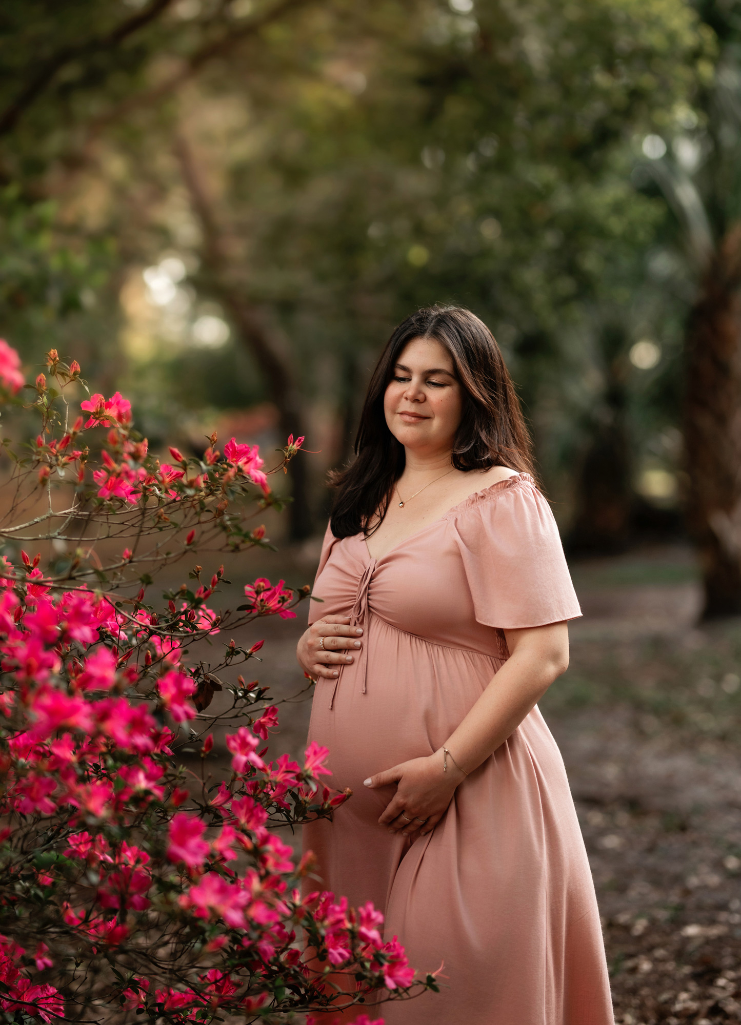 Pregnant woman in pink dress standing outdoors near pink flowers, smiling with eyes closed, surrounded by trees.