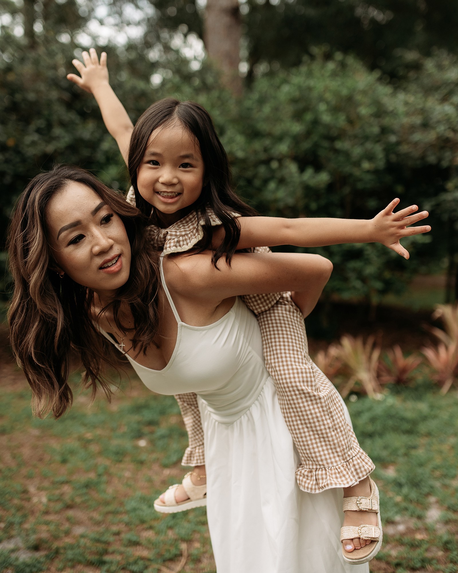 A woman carrying a smiling young girl on her back outdoors with trees and grass in the background.