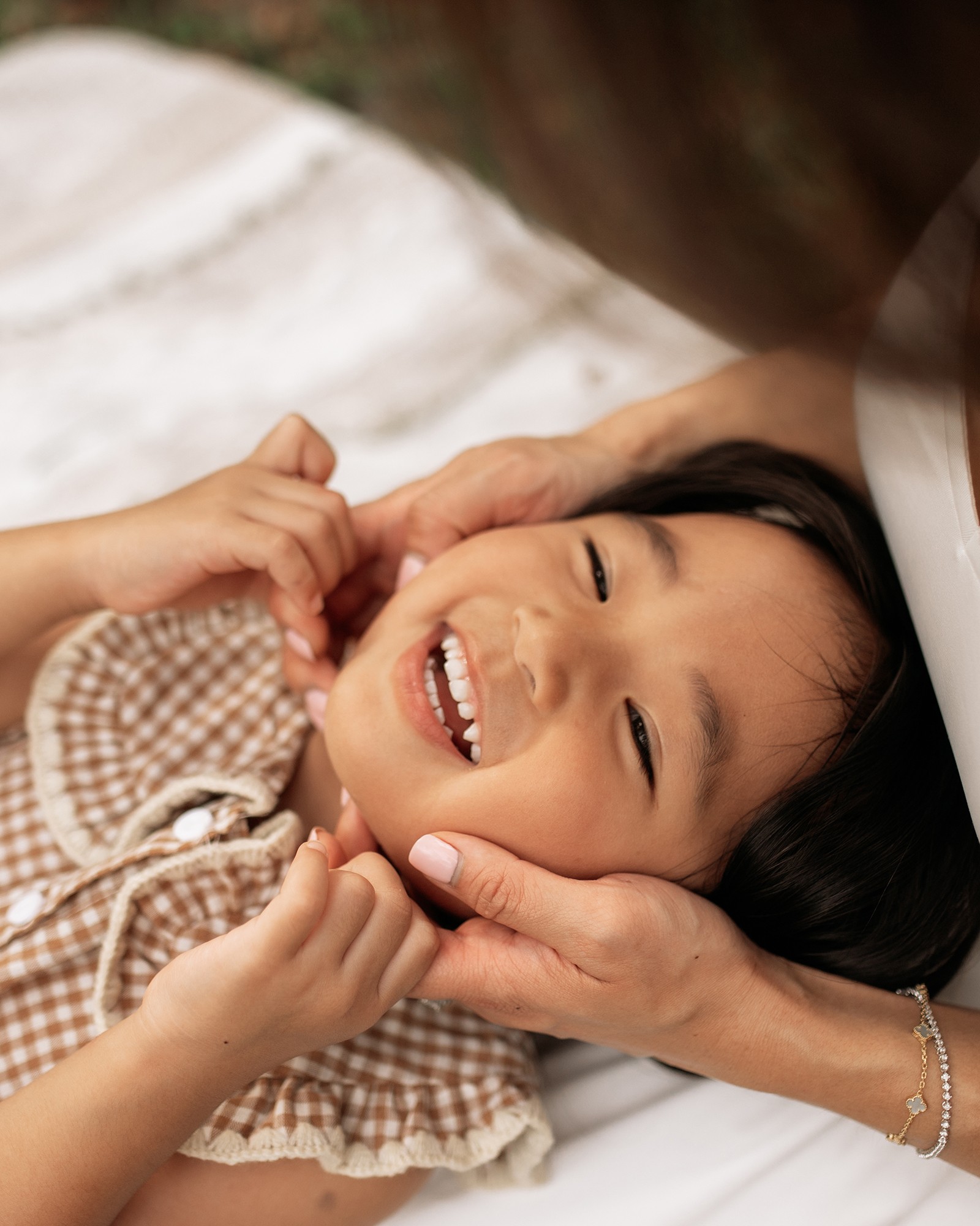 Child lying down, smiling and laughing, being gently held by an adult's hands, with a blurred background.