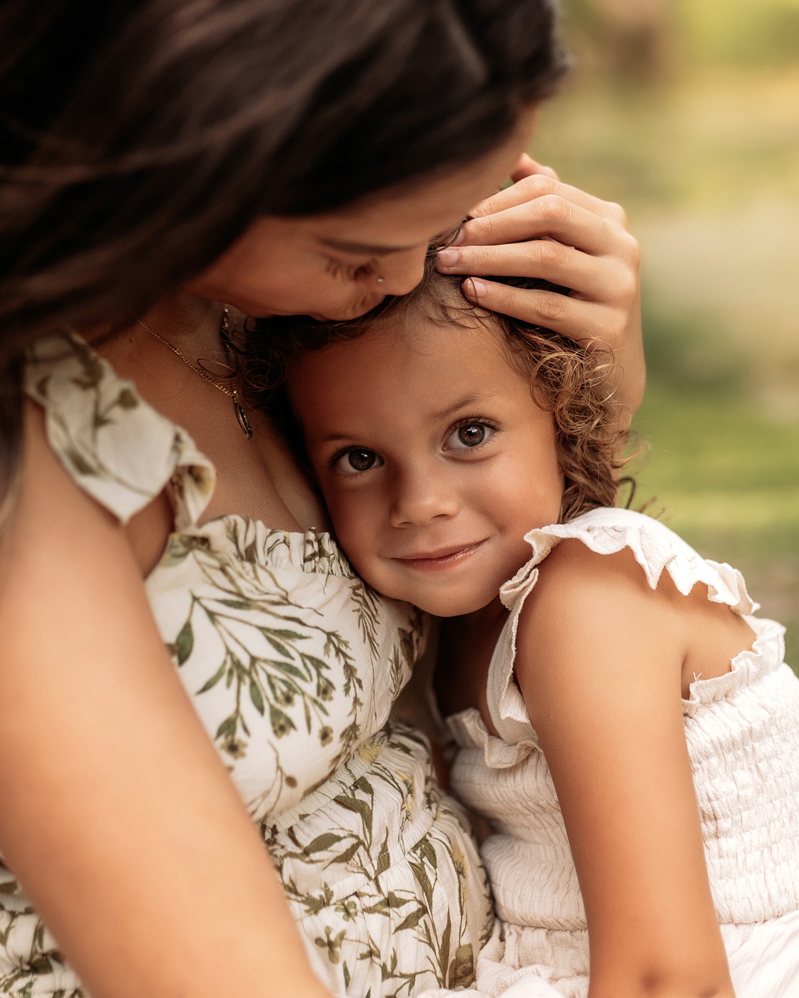 Adult holding a young girl, both smiling, outdoors with greenery in background.