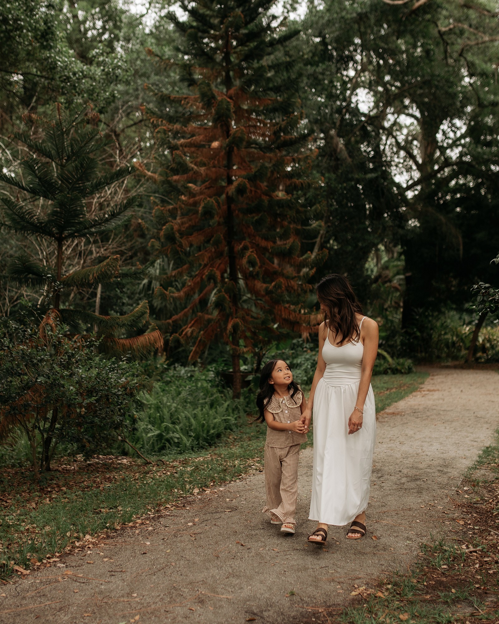 Two women walking on a dirt path in a forested area, surrounded by trees and greenery.
