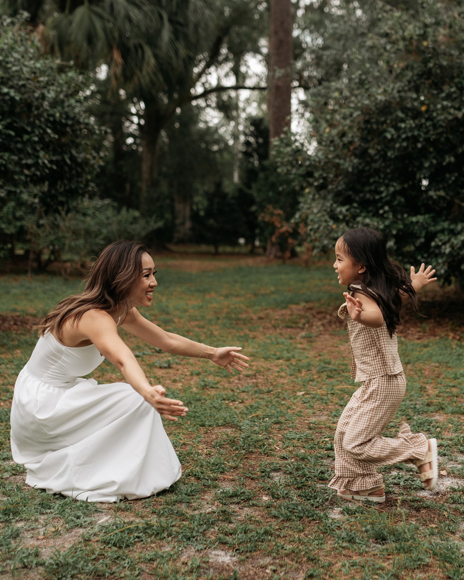 Two girls playing outdoors on grass, one in a white dress crouching and the other in beige pants with arms raised.