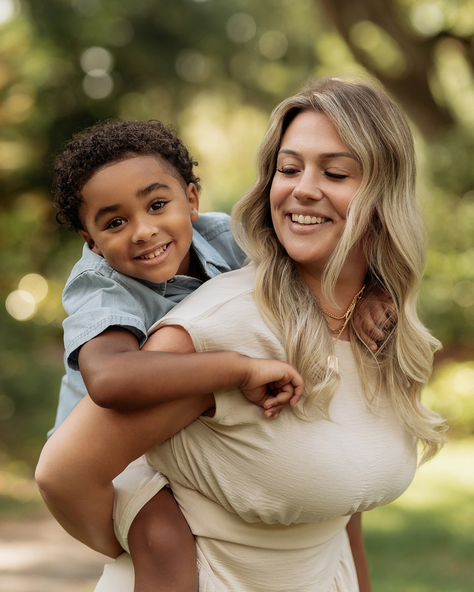 Woman carrying a smiling child on her back outdoors with trees in the background.