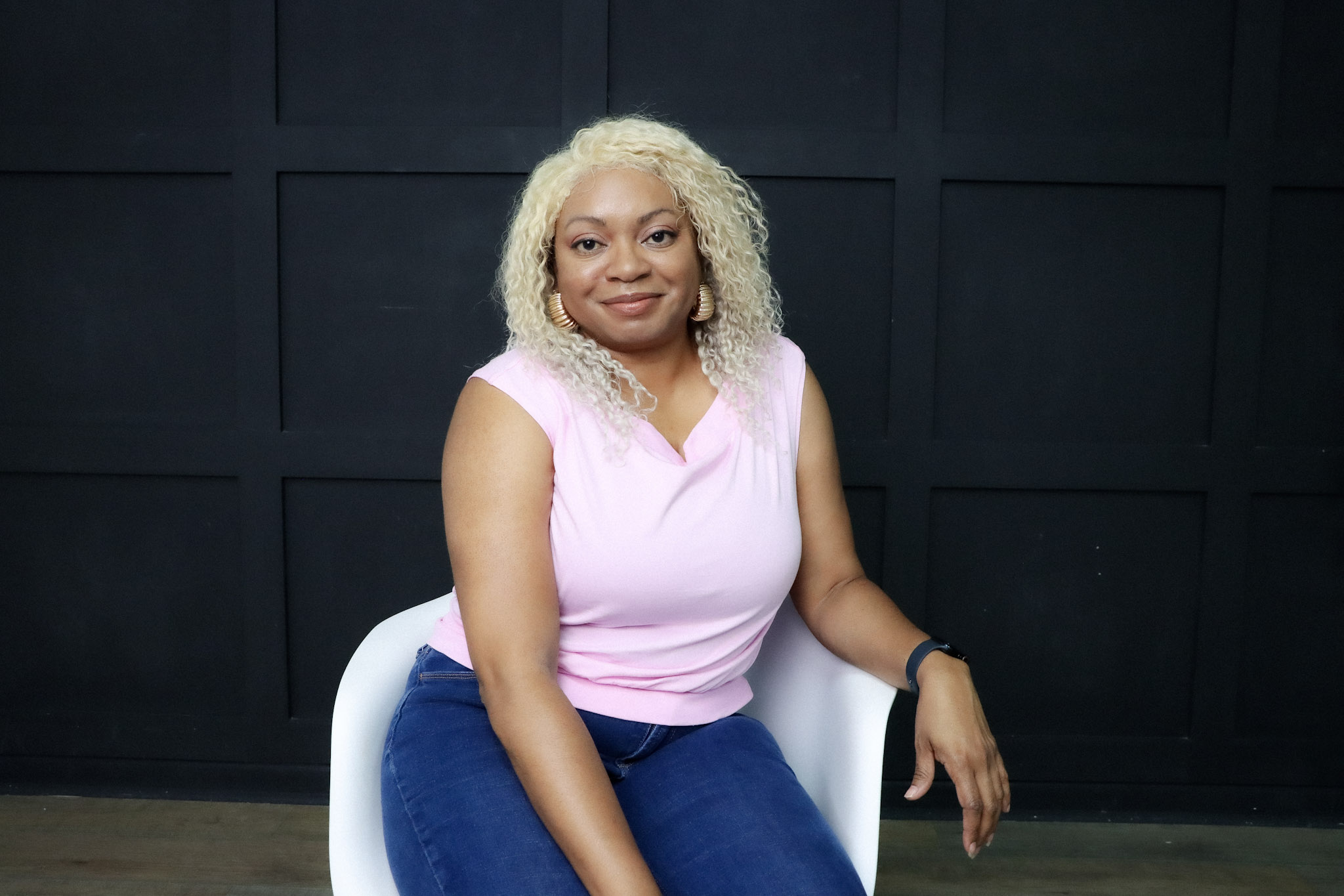 Woman with blonde curly hair sitting on a white chair against a dark wall, smiling, wearing a pink top and blue jeans.