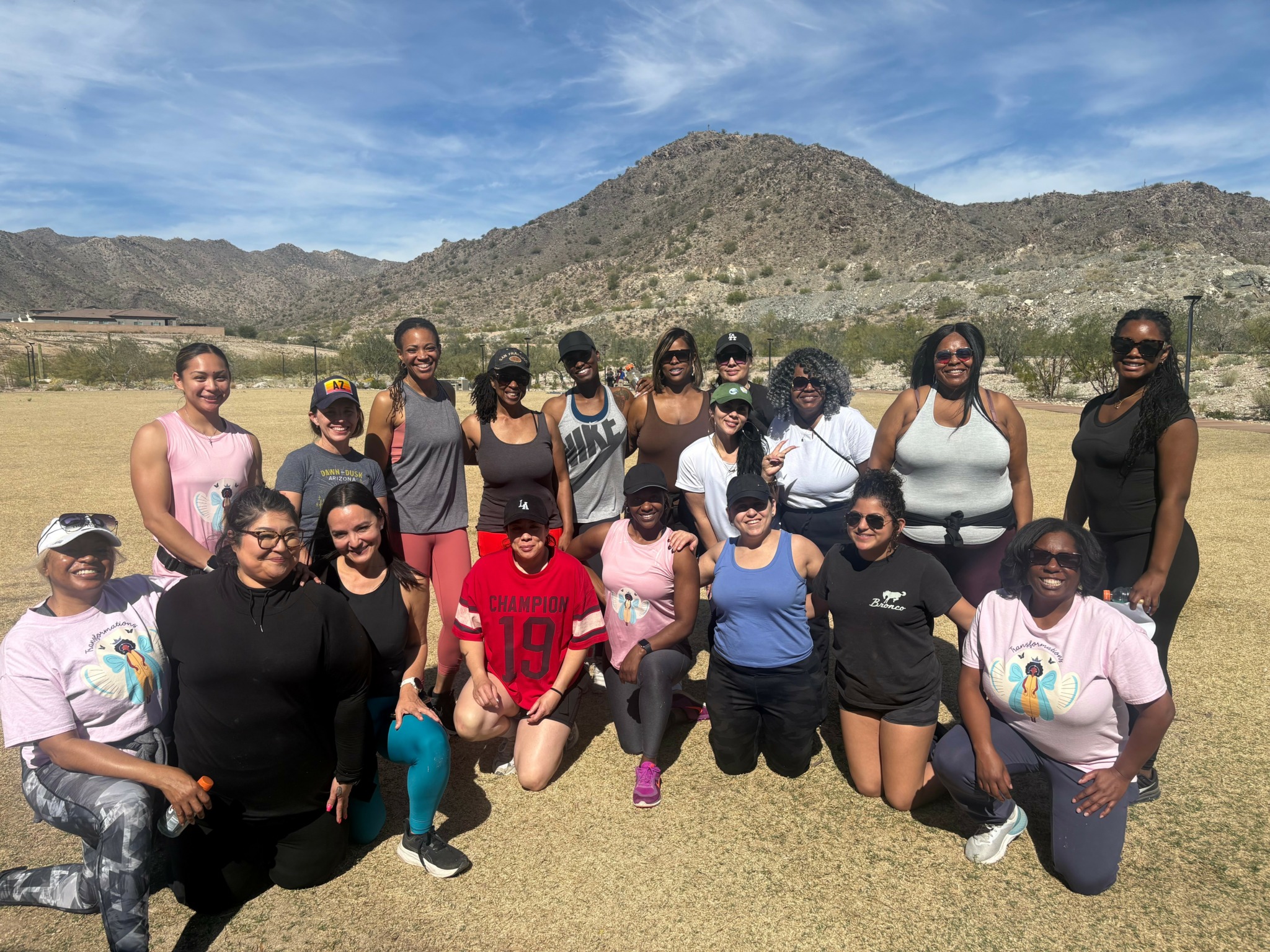 Group of 15 people outdoors with mountains and blue sky in background, some wearing sunglasses and casual clothing.
