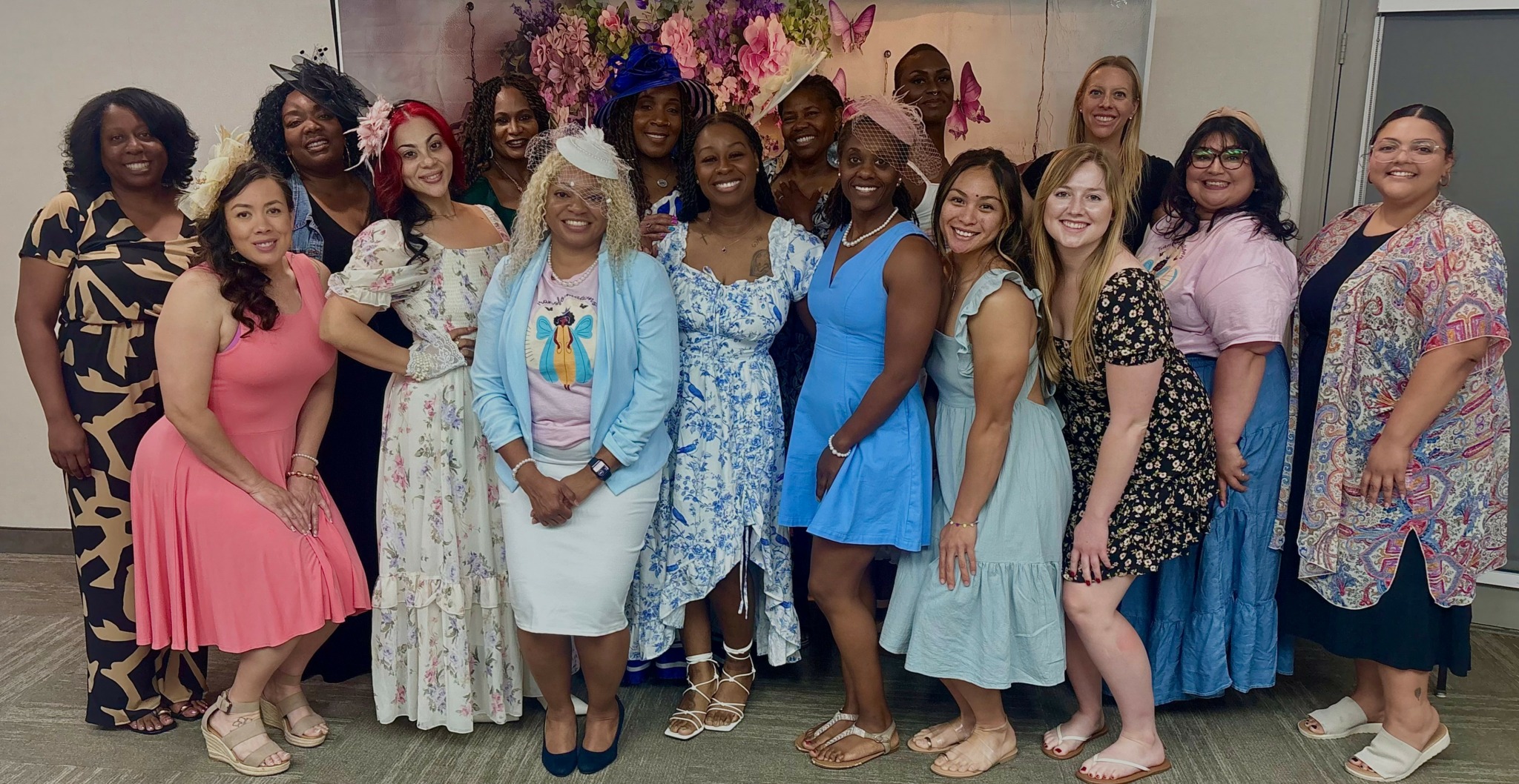 Group of women standing together indoors, smiling, with colorful flowers in the background.