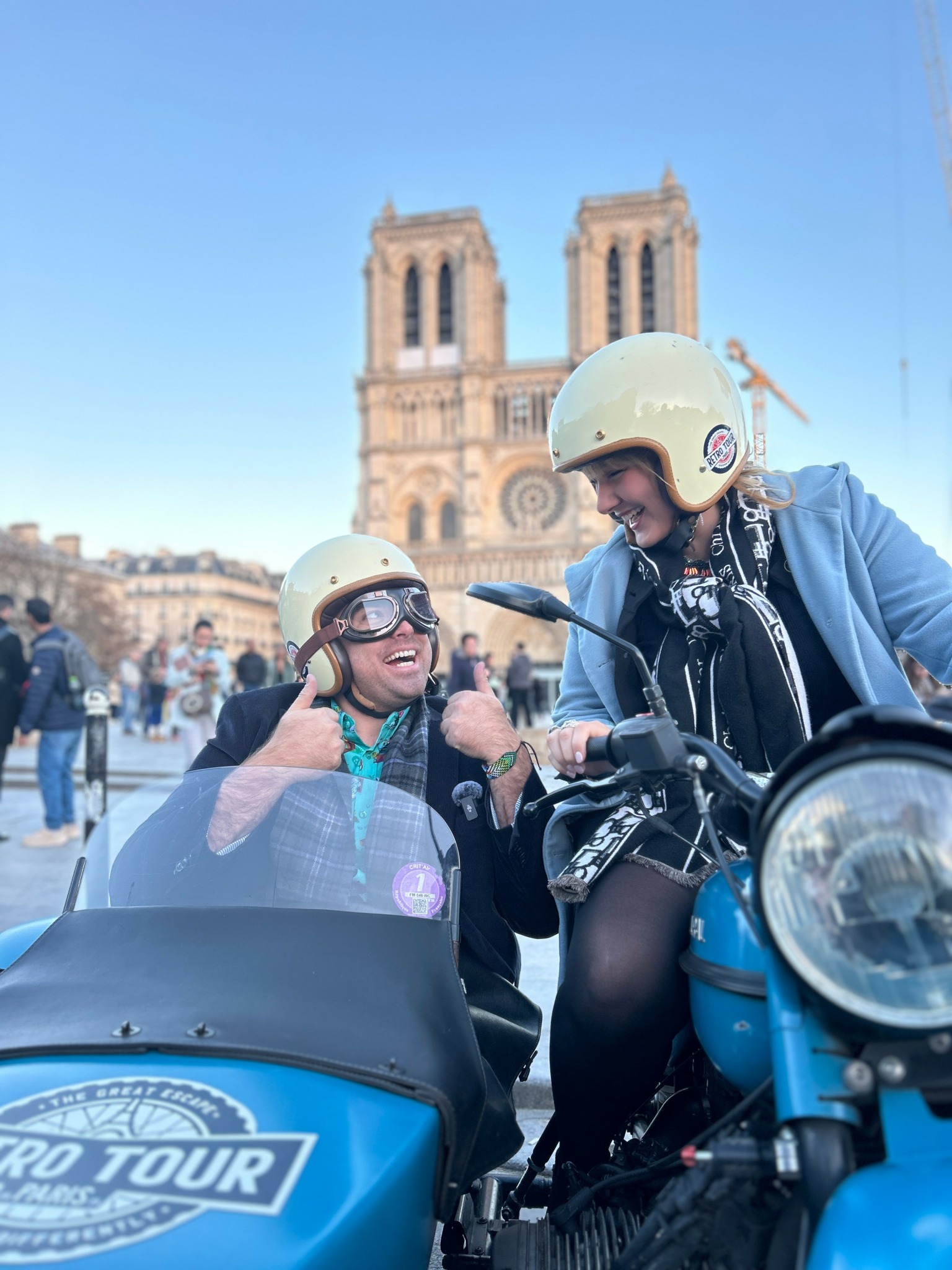 Two women wearing helmets sit on a motorcycle in front of Notre-Dame Cathedral, smiling and talking, with a clear blue sky.