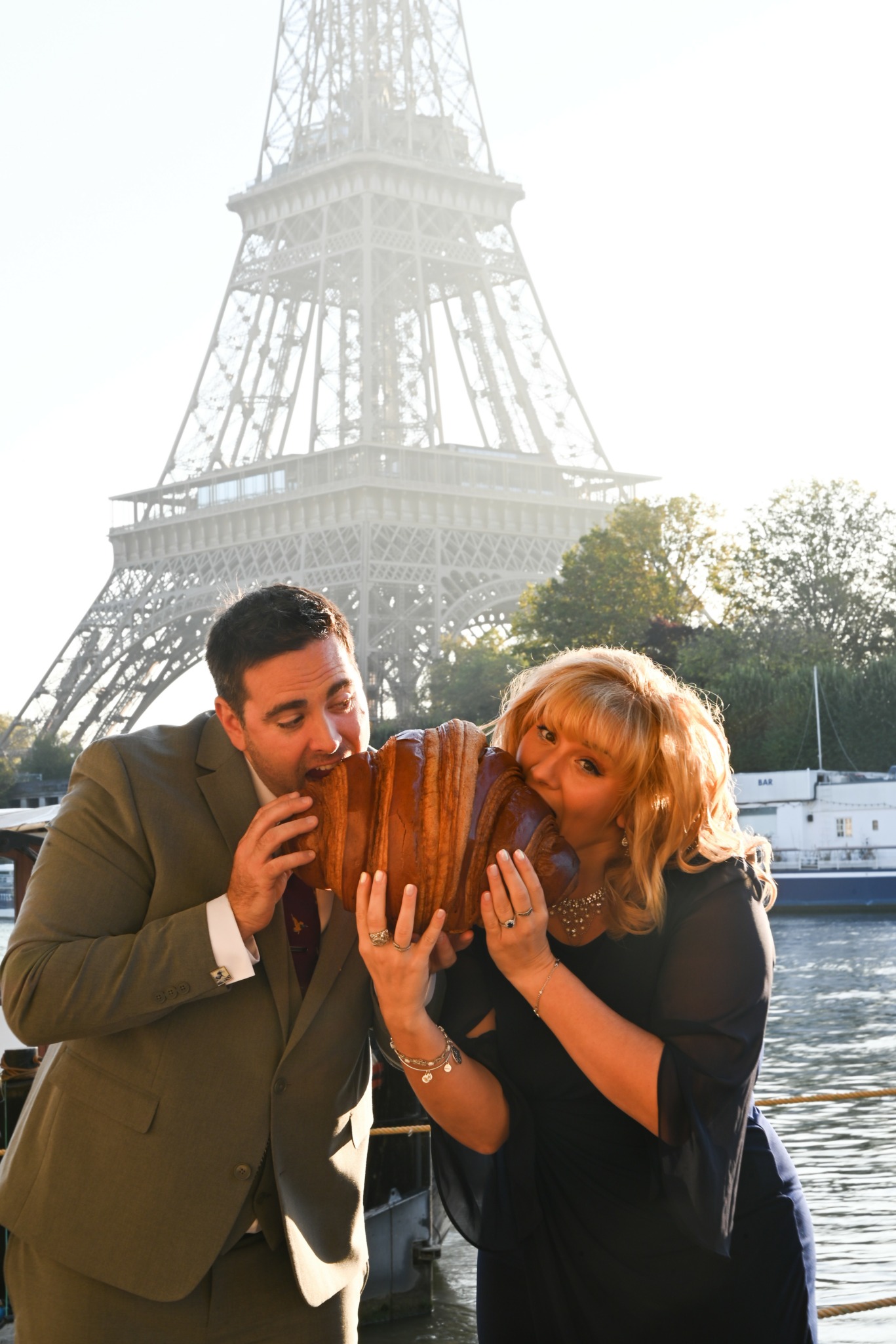 Two people, a man and a woman, hold a large loaf of bread near a river with the Eiffel Tower in the background.