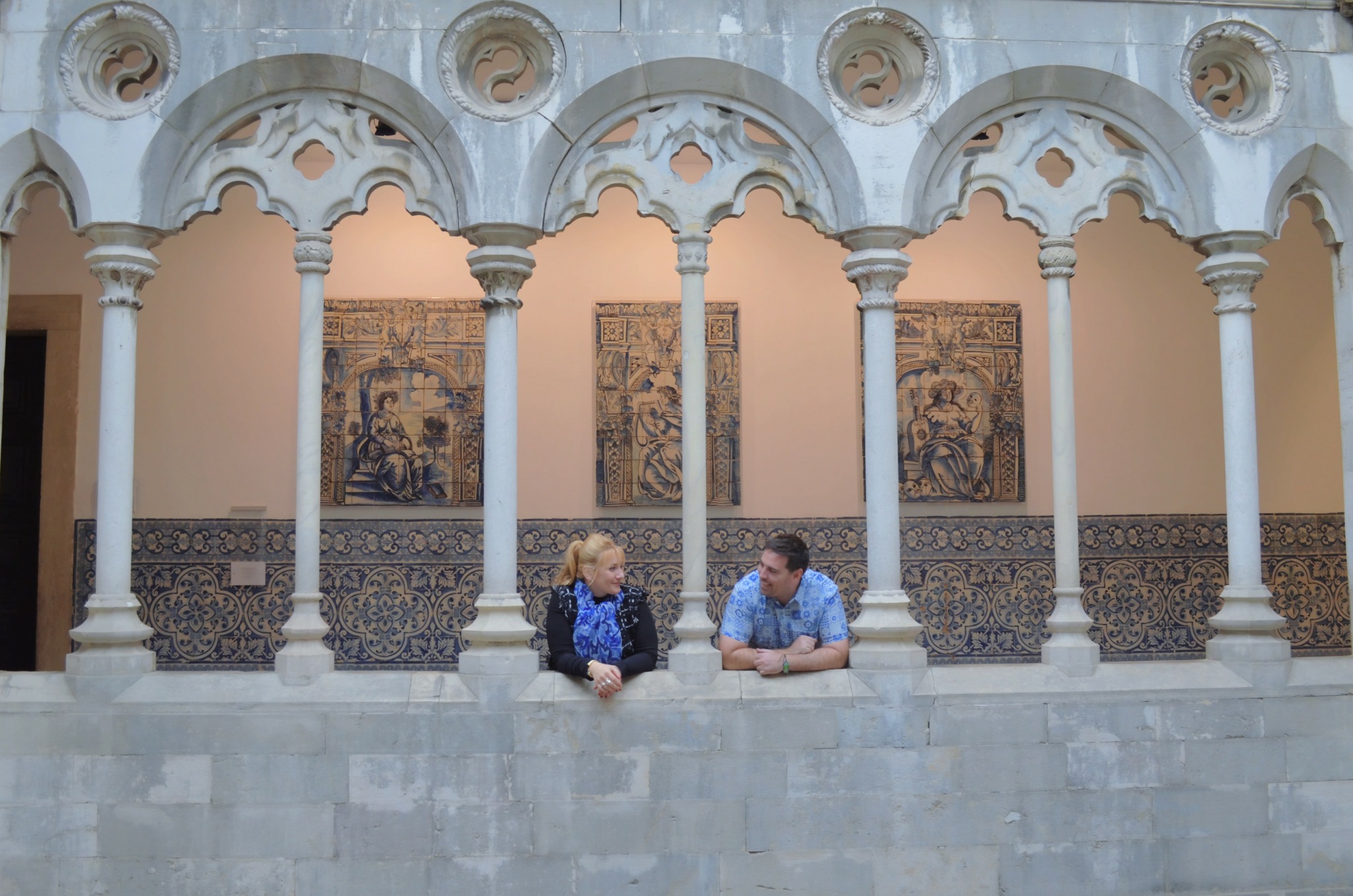 Two people sit at a marble ledge in front of ornate arches and wall art, with decorative tiles behind them.