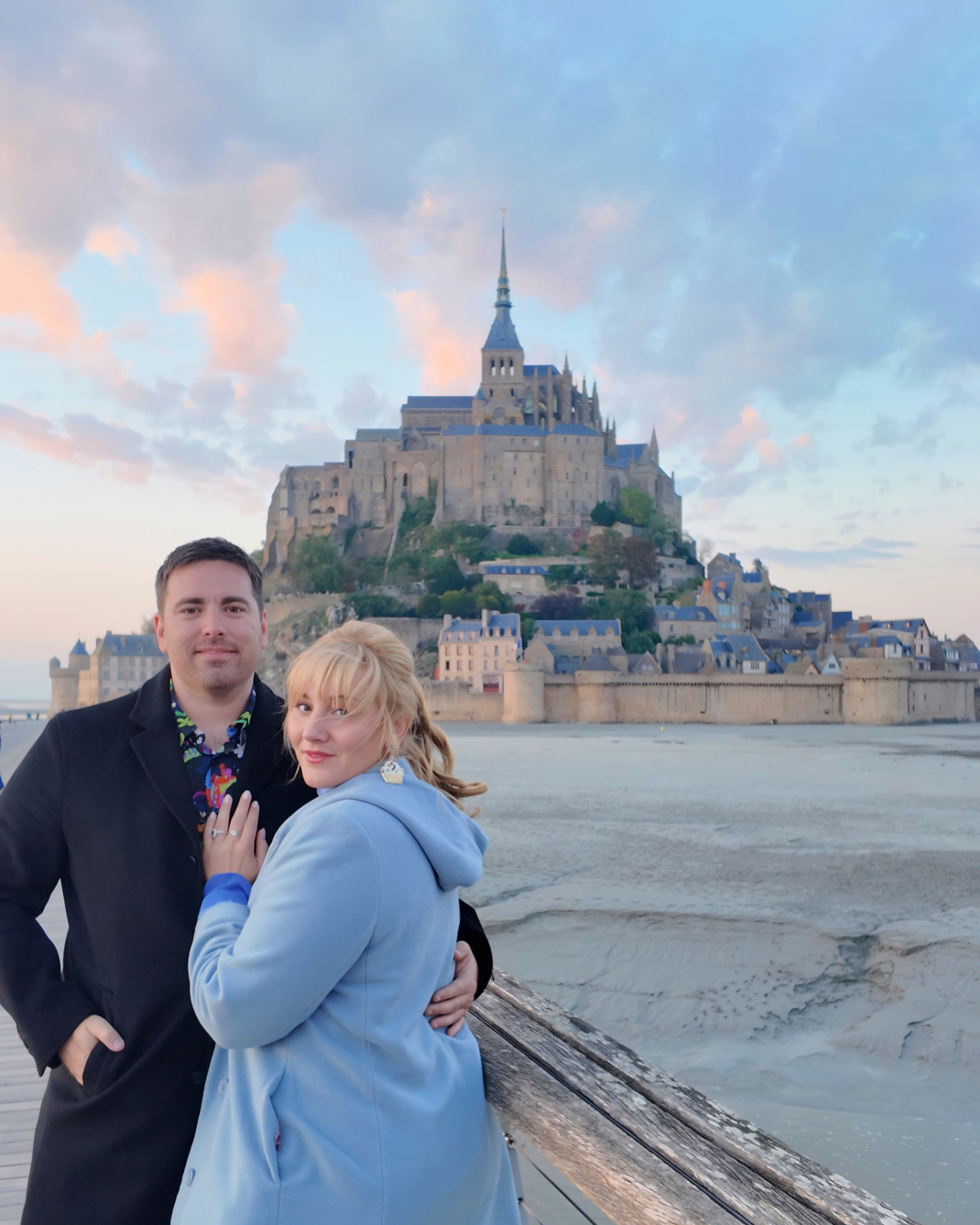 A man and woman stand together in front of Mont Saint-Michel, with the island and abbey in the background during sunset.