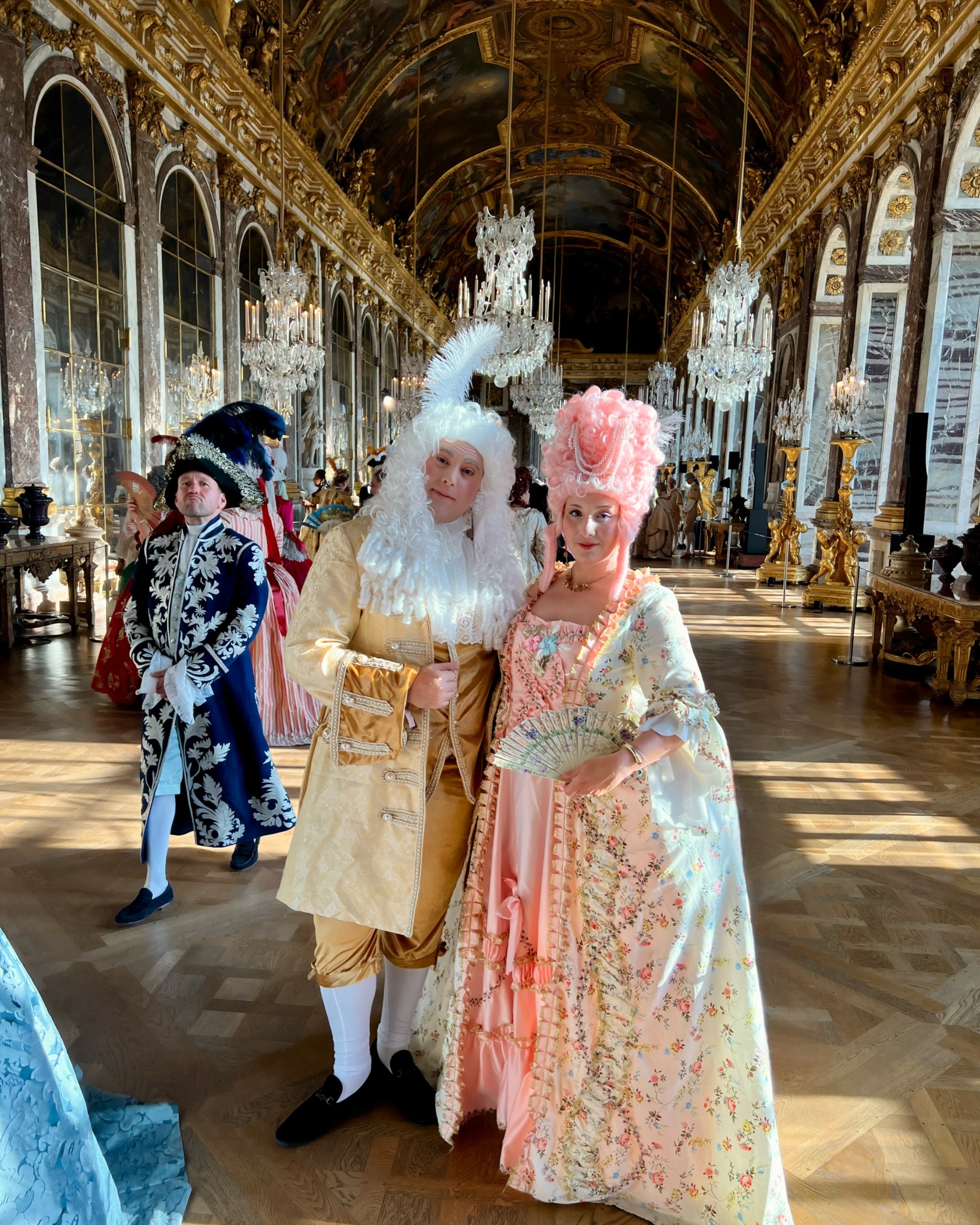 Two people in elaborate 18th-century costumes pose in a grand, ornate hall with chandeliers and gilded decorations.