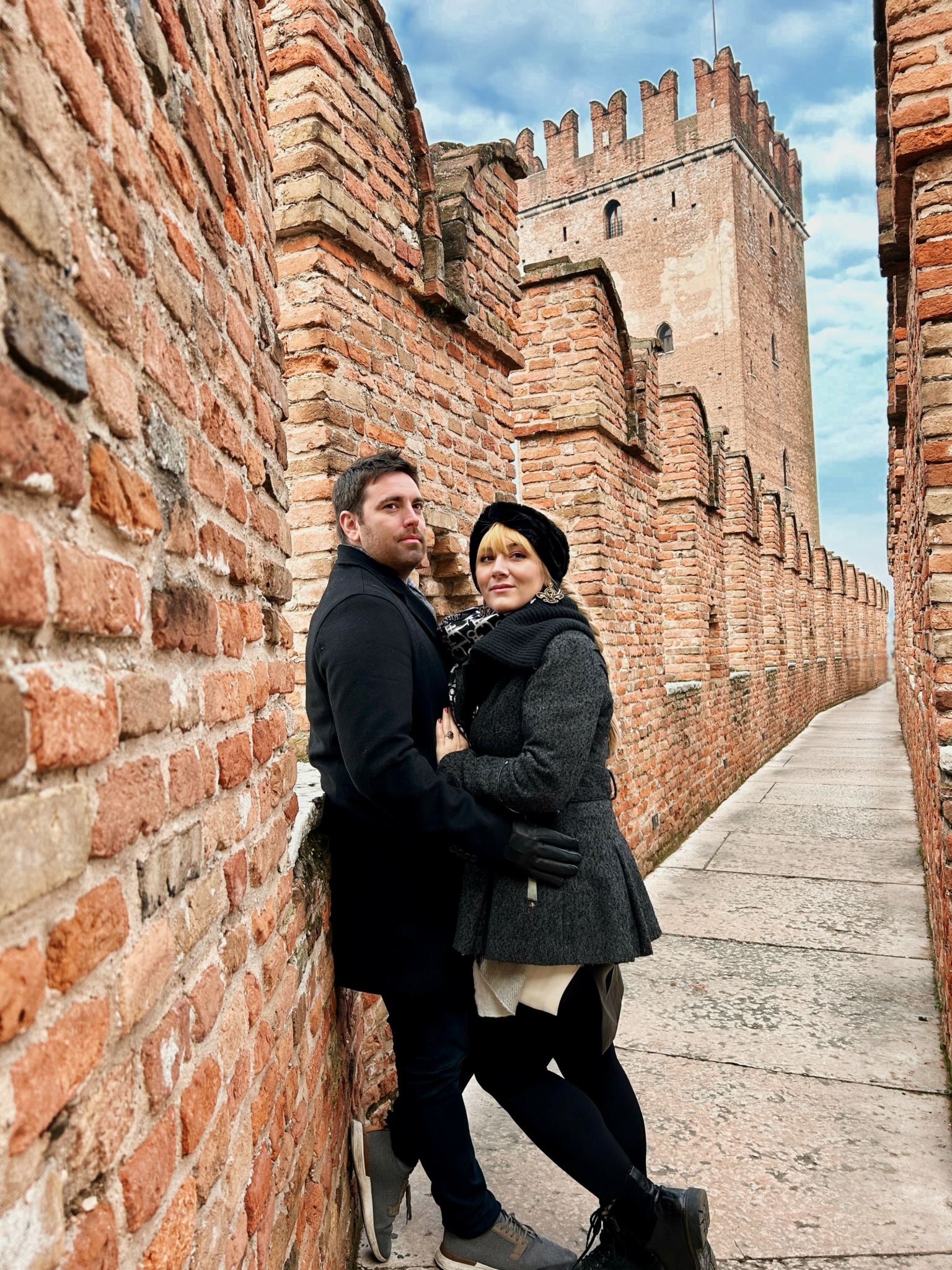 A man and woman stand close together in a narrow brick alley with a castle tower in the background.