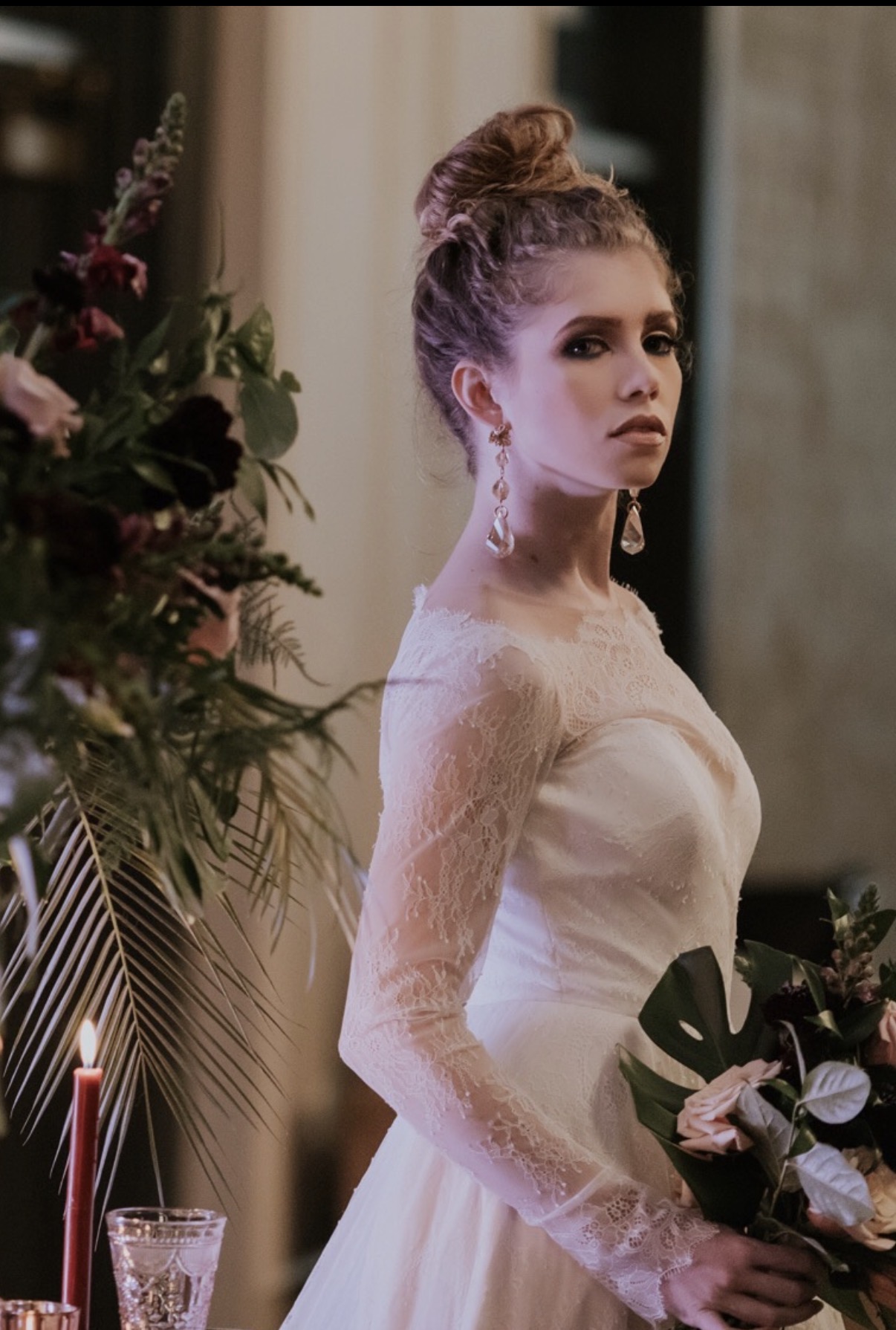Young woman in a white lace dress with an updo hairstyle holding a bouquet, standing near flowers indoors.