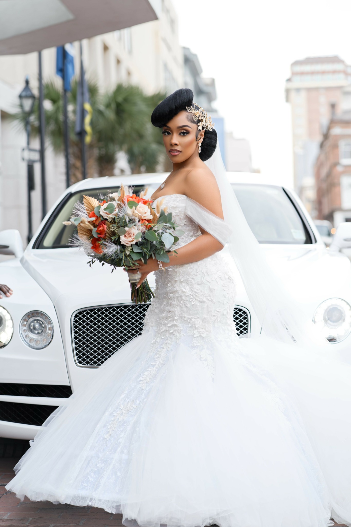 Bride in wedding dress holding a bouquet, standing in front of a white luxury car on city street.