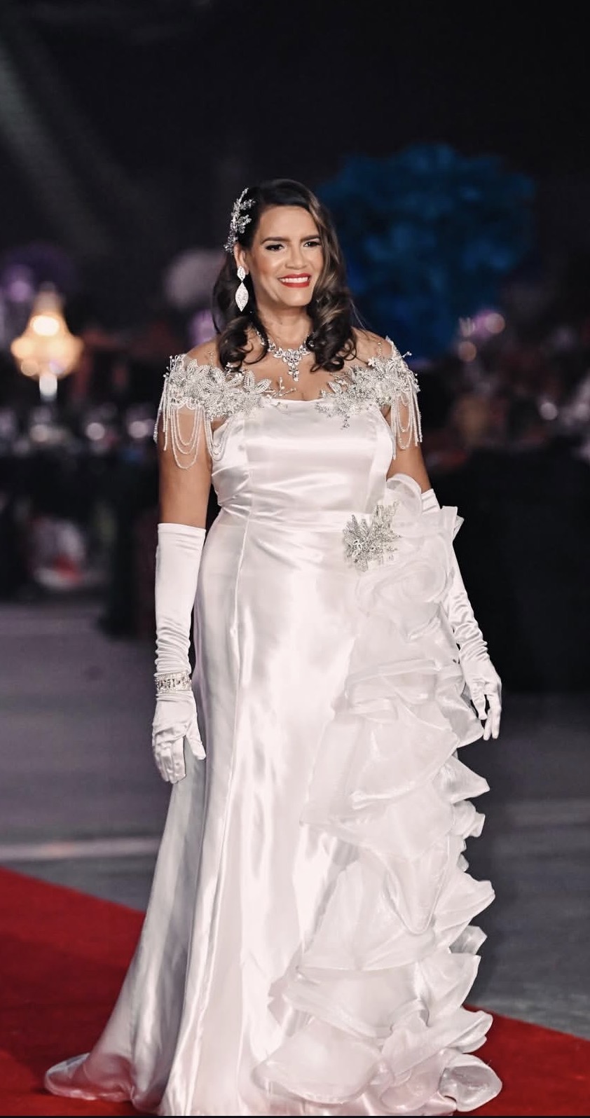 Woman in a white wedding dress with lace details, standing on a red carpet at night, smiling, with a dark background.