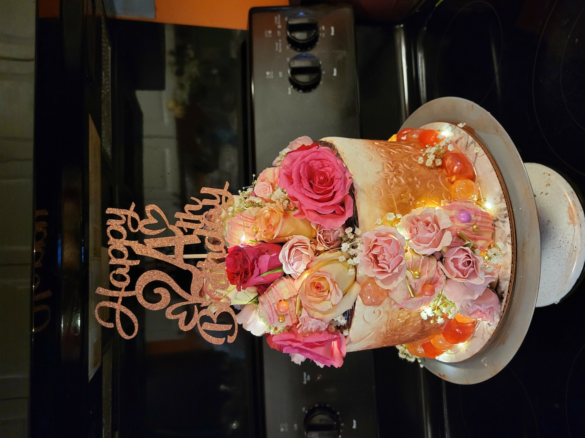 A birthday cake decorated with pink and peach roses, with a 'Happy Birthday' topper, on a table with a TV and electronic devices in the background.