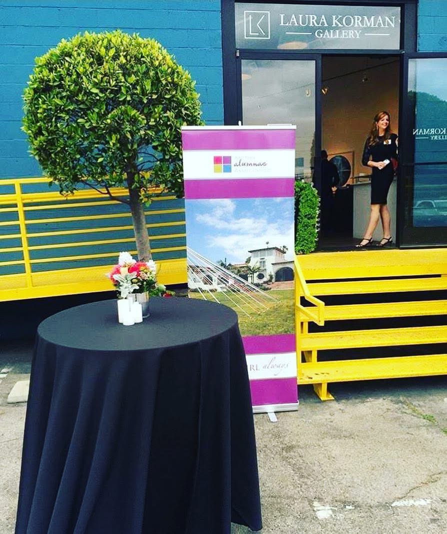 A round table with a black tablecloth and a flower arrangement, outside an art gallery with a woman in black, a tree, and a sign. Word count: 20