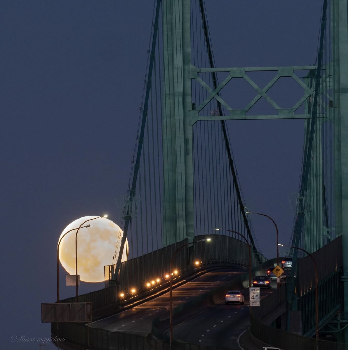 Full moon near bridge structure with vehicles on the road, night scene, illuminated streetlights, dark sky.