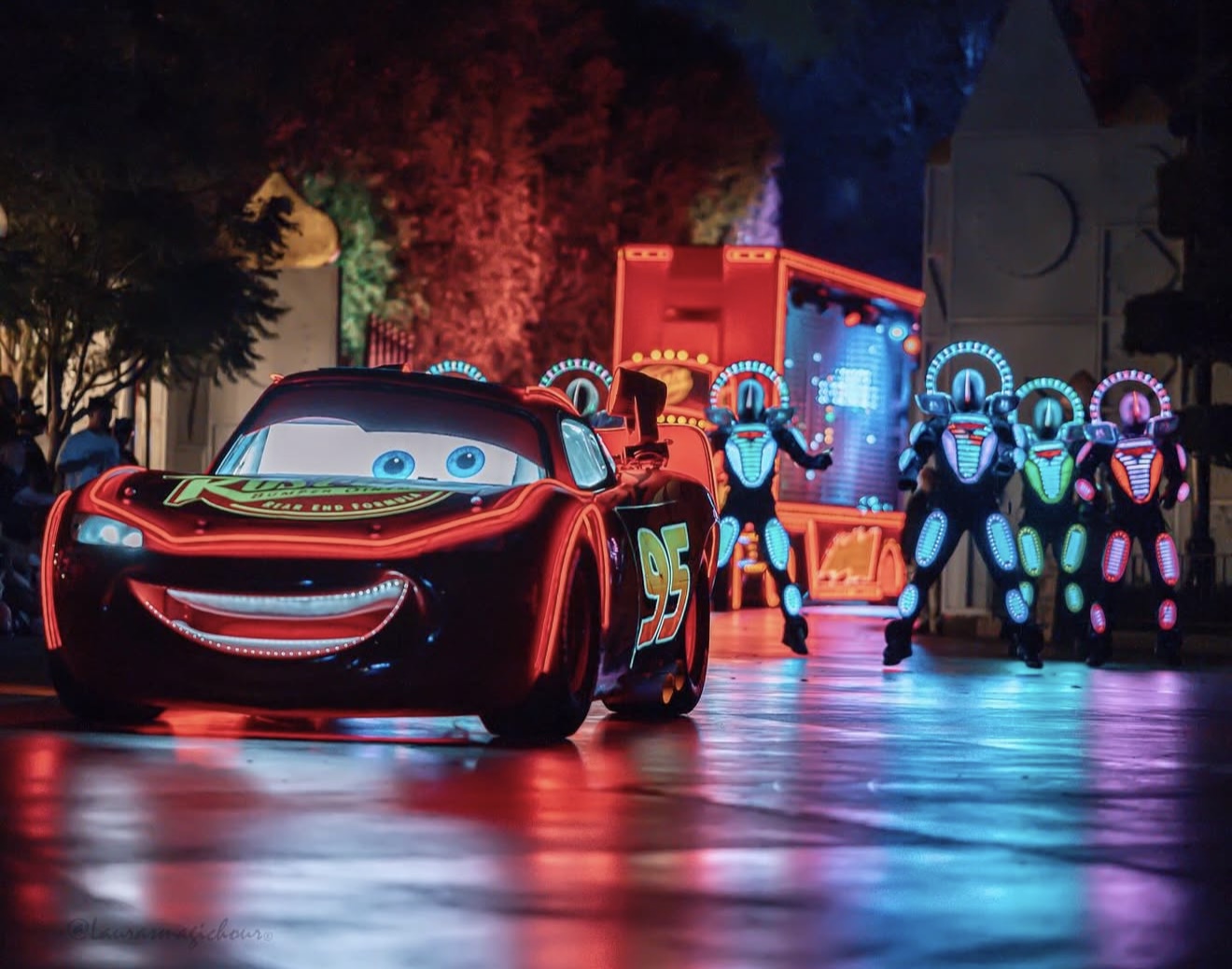 Animated race car character with a big smile and eyes, illuminated by colorful lights at night, with other characters in the background.